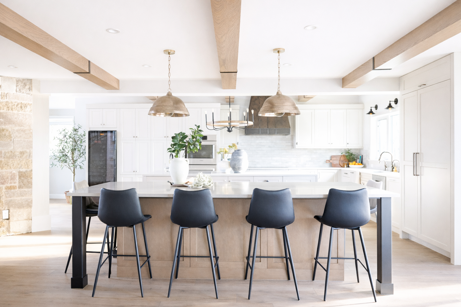 Bright, modern kitchen with white cabinets, a large island with a beige base and black legs, and five black bar stools. Decor includes large white vases with green plants, a wooden cutting board, and yellow flowers. Overhead lighting features two met