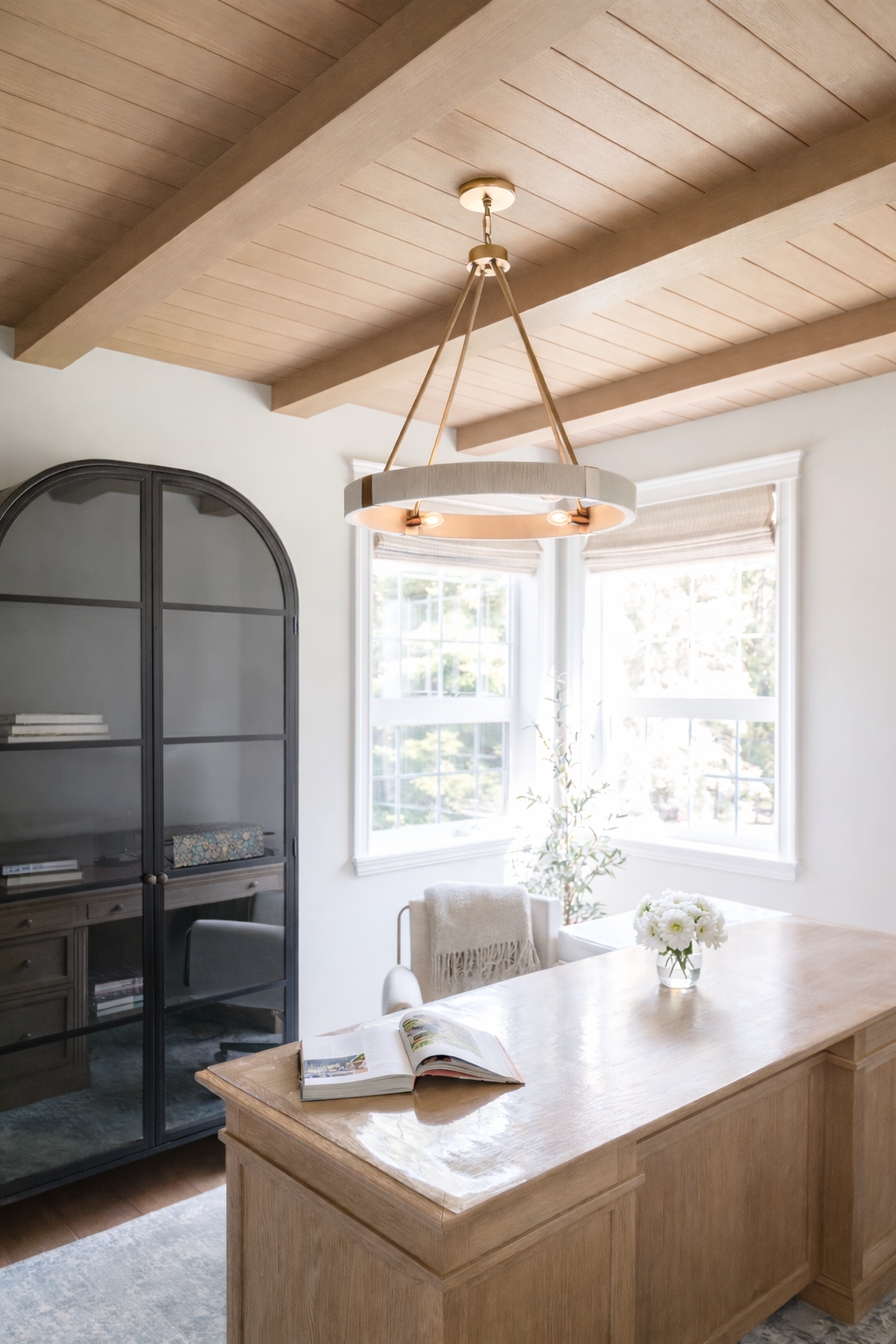 A dining room with a wooden table, a black glass-front cabinet, and large windows letting in natural light, decorated with a vase of white flowers.