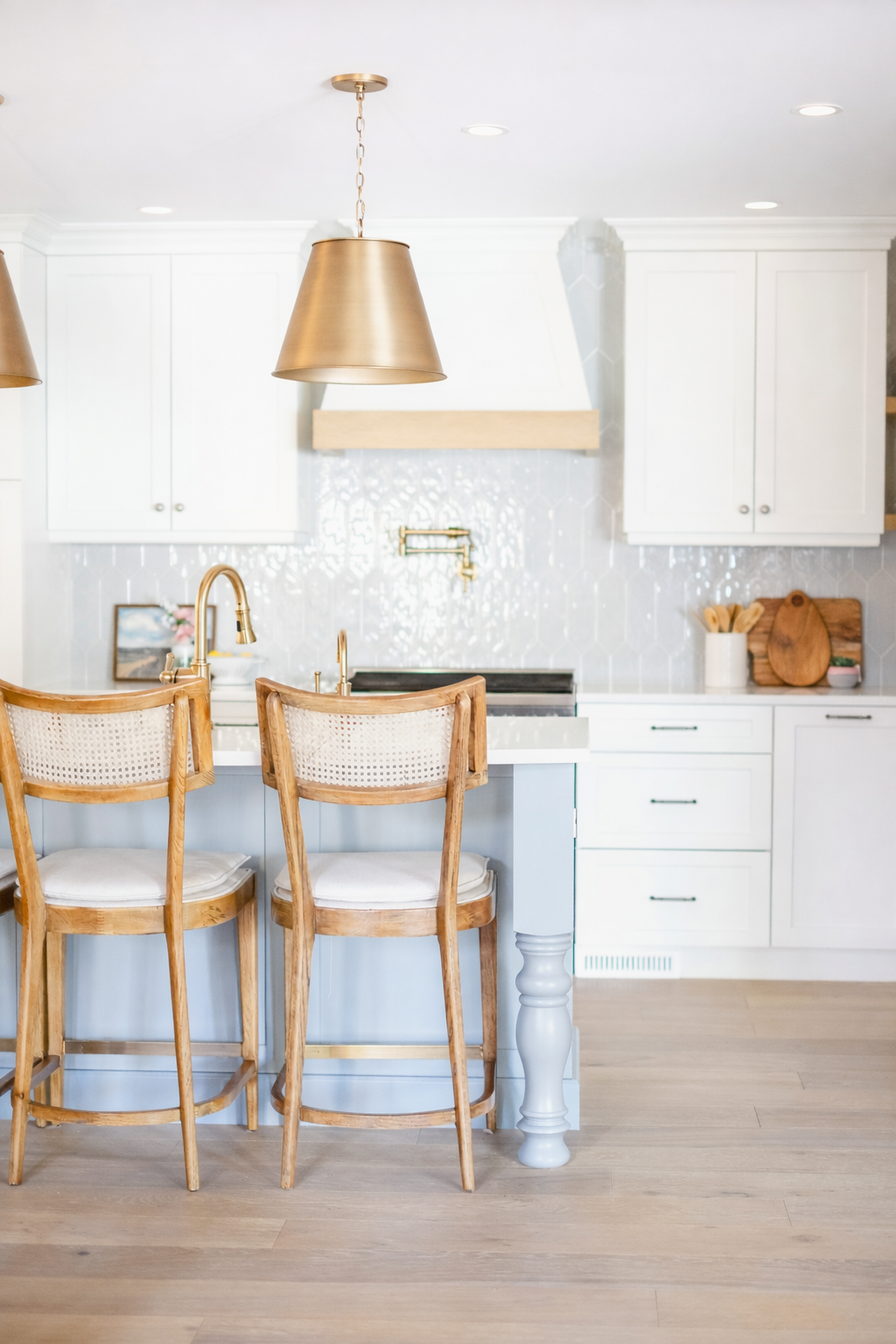 A bright kitchen with white cabinets, a white tiled backsplash, and a white countertop with a sink. There are two wooden barstools with white cushions at the kitchen island, which has a light blue-painted front. Gold pendant lights hang above the isl