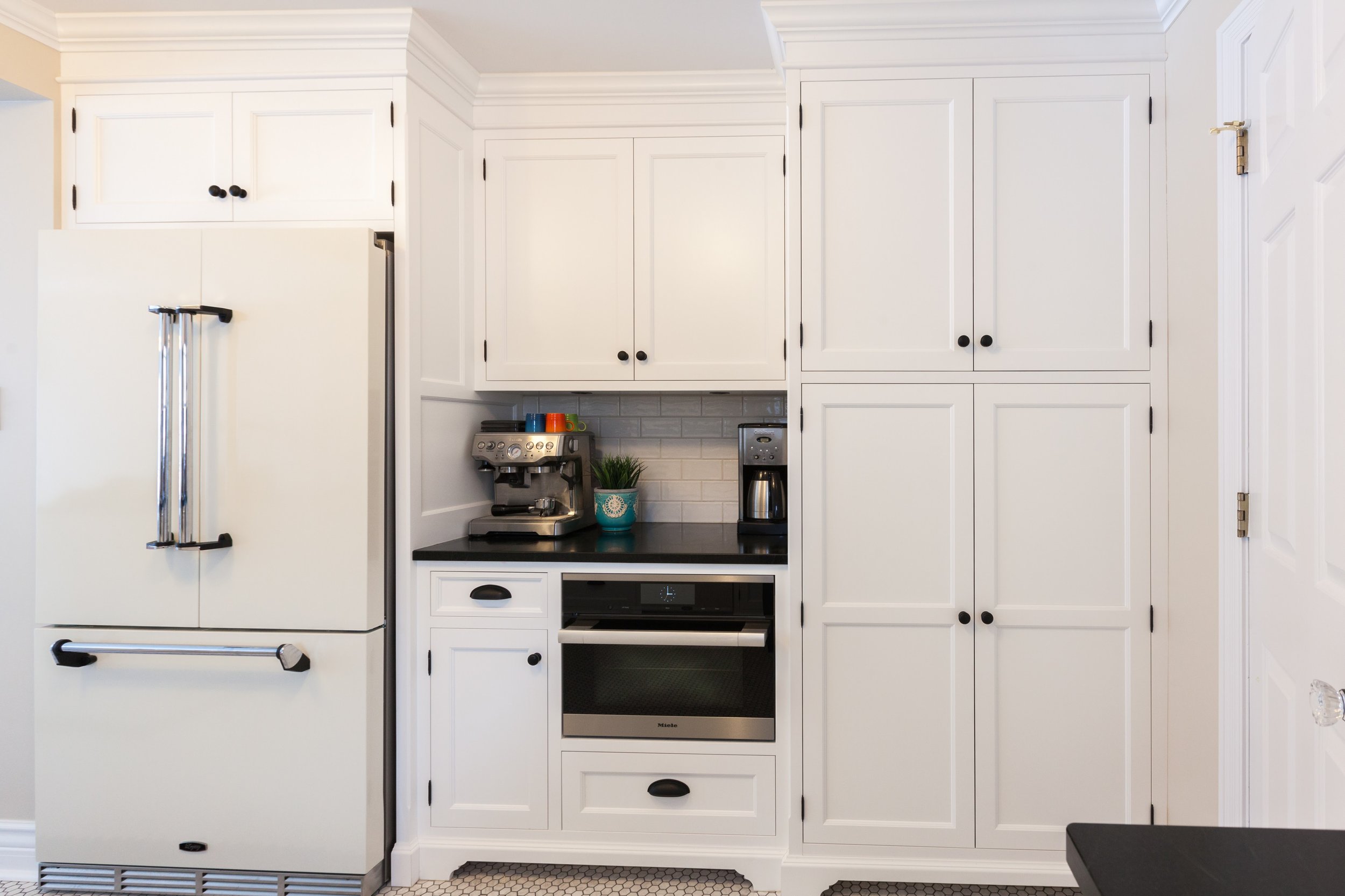 White kitchen with black countertops, a refrigerator, a coffee maker, a espresso machine, upper and lower cabinets, and a small open counter space with strawberries and a potted plant on the counter.