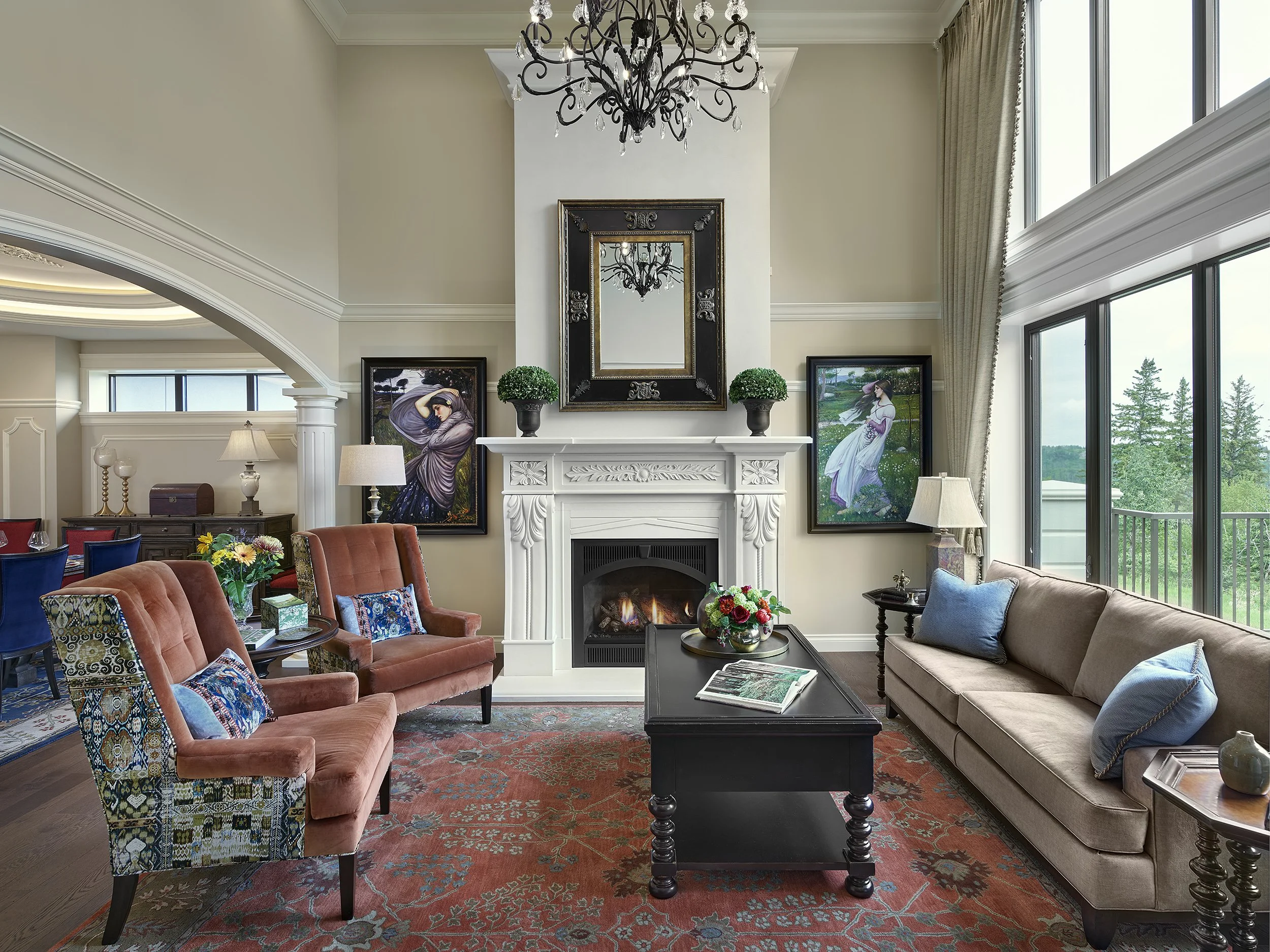 Living room with fireplace, large windows, and elegant furniture including beige sofa and pink armchairs, decorated with paintings, potted plants, and flowers.