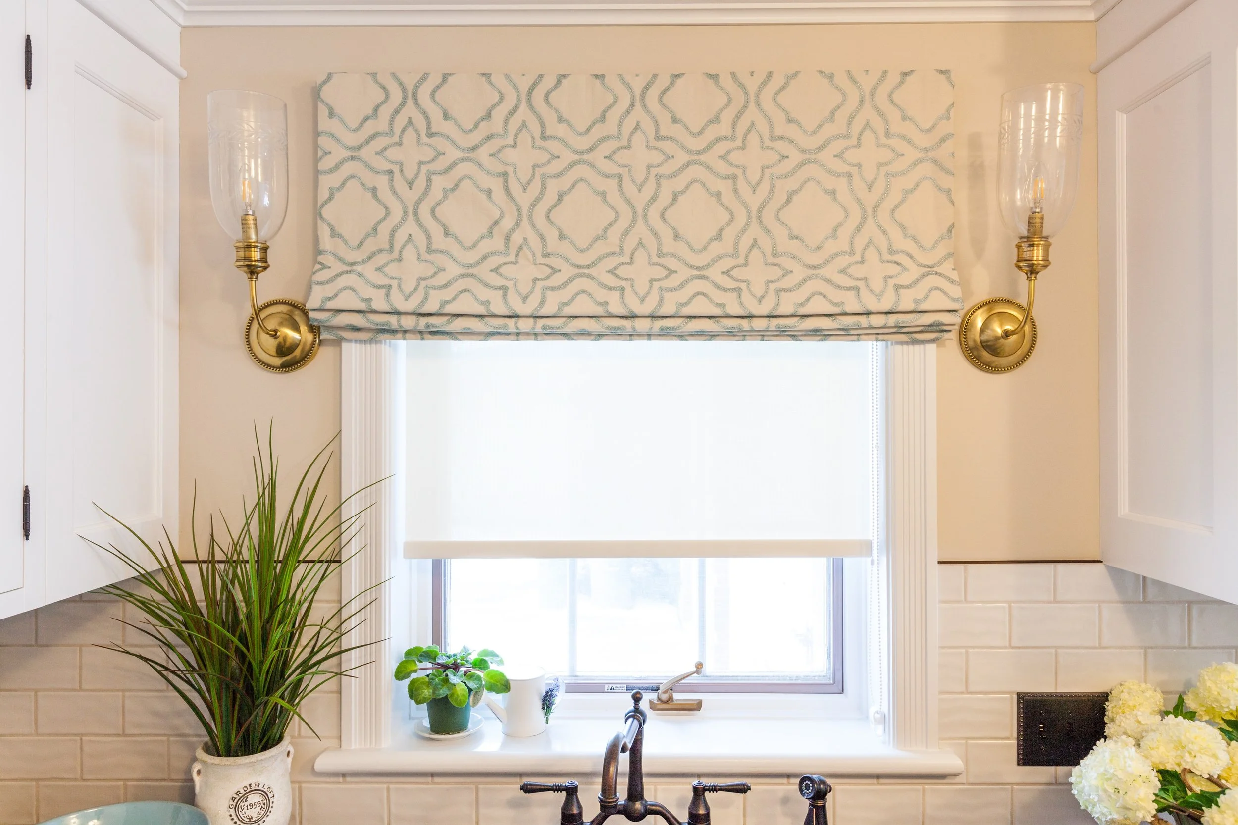 Kitchen window with white frame, featuring a patterned valance and roman shade, surrounded by white cabinetry and wall-mounted antique brass sconce lights, with potted plants and hydrangeas on the windowsill.