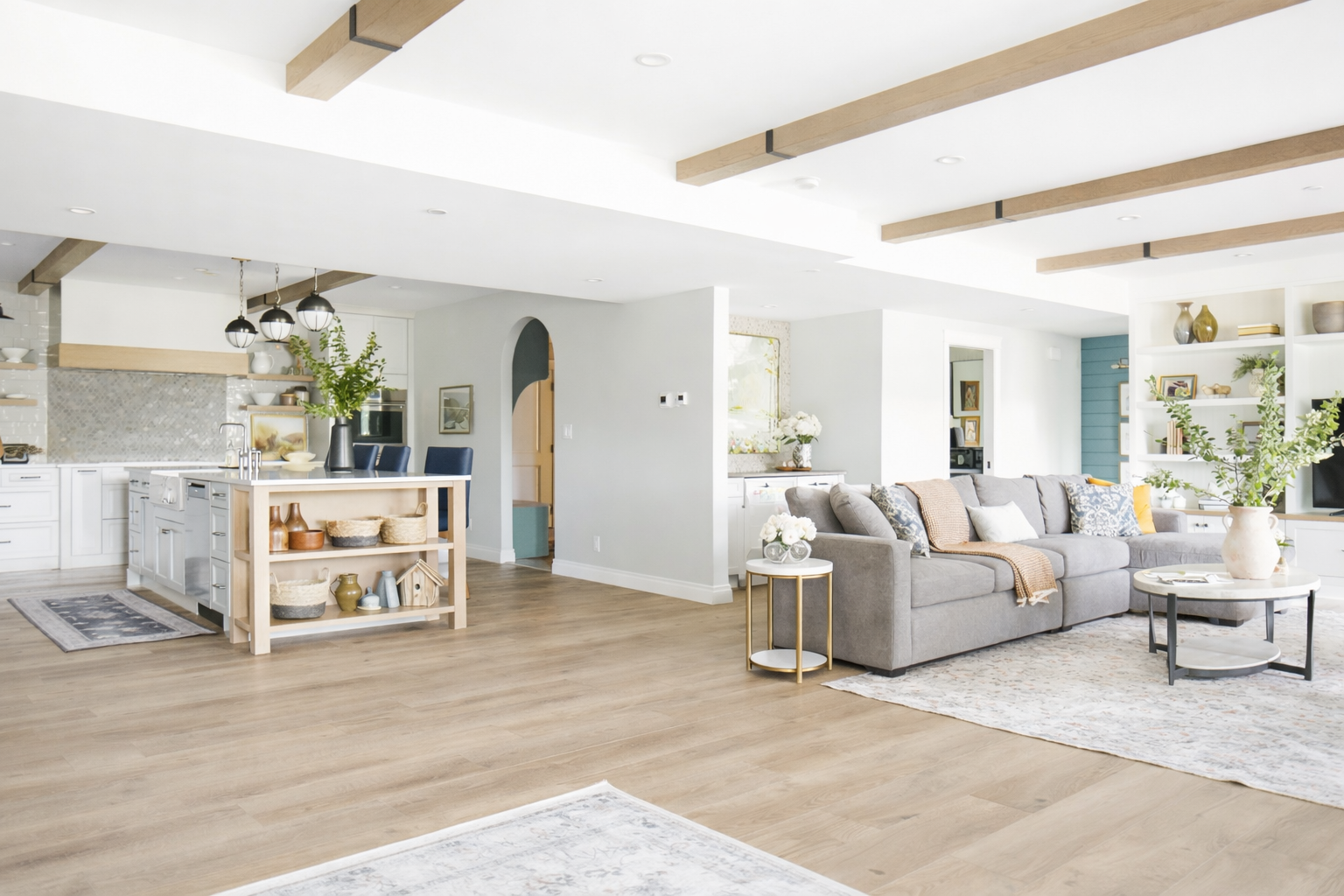 Bright, open-concept living room and kitchen with wood beams, white walls, and light wood flooring.