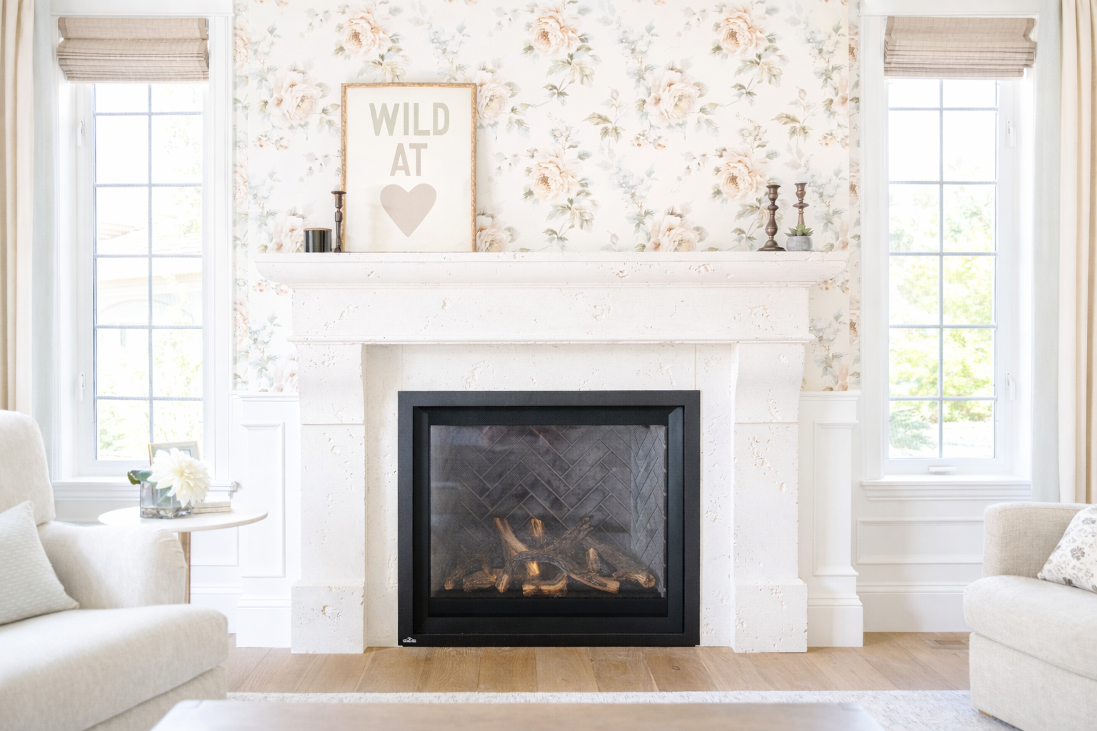 A cozy living room with a white marble fireplace, floral wallpaper, and two windows with beige shades. Decorations include a framed sign reading 'WILD AT ♥', candlesticks, small potted plants, a vase with white flowers, and couches with cushions.