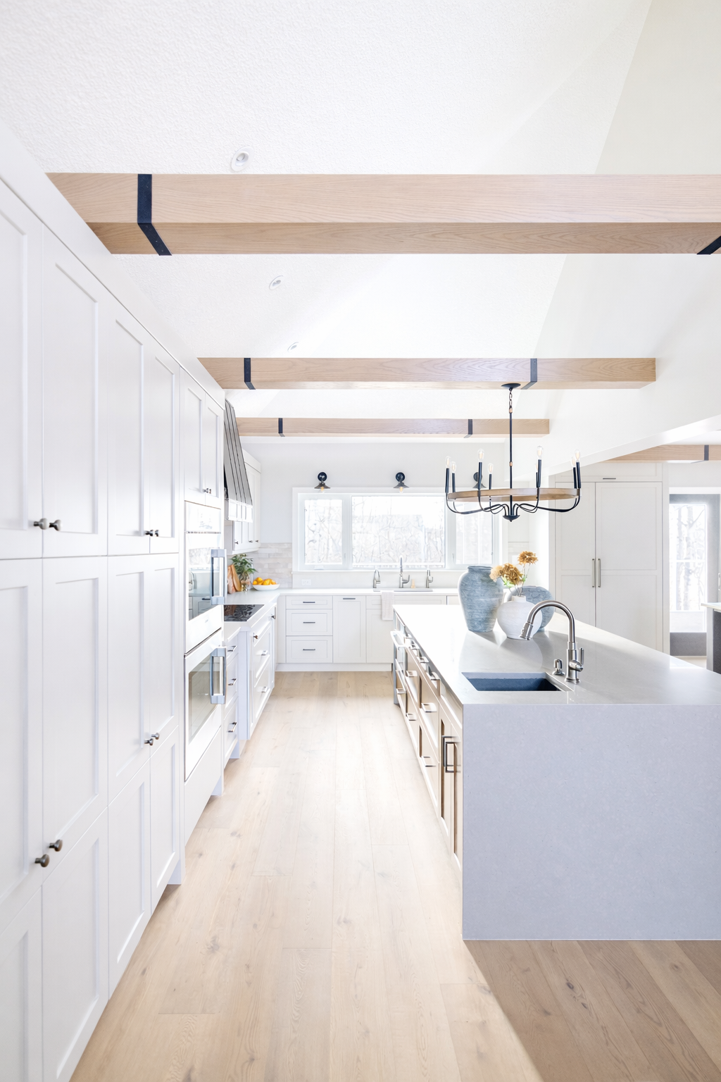 Bright modern kitchen with white cabinets, light wood flooring, a central island, and decorative wood beams on the ceiling.