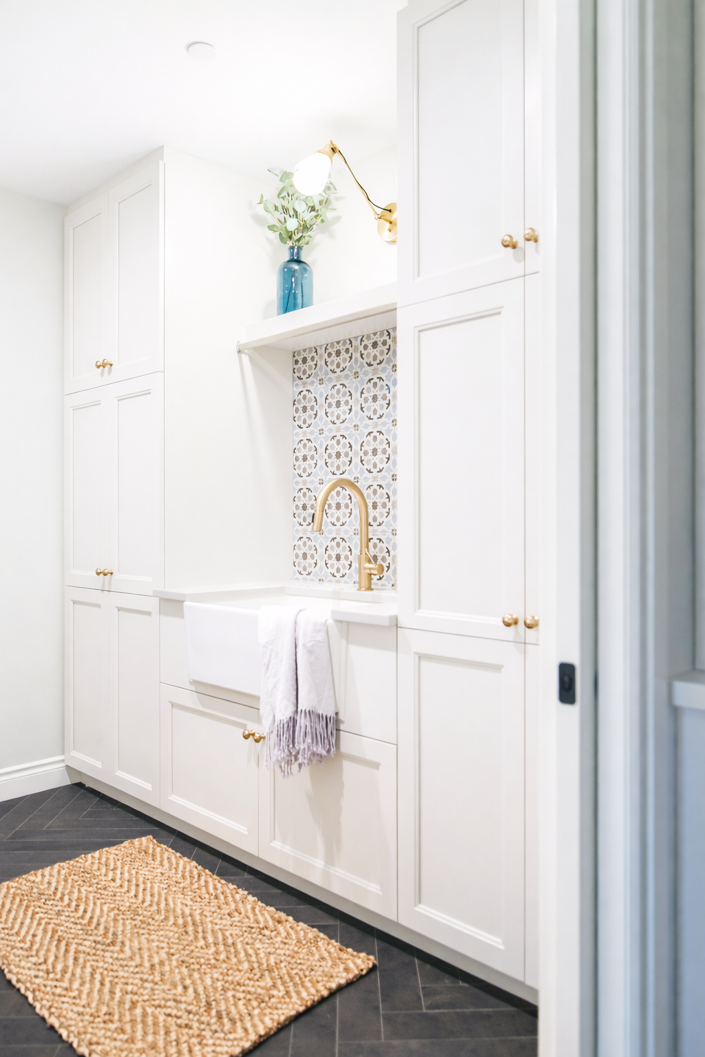 A bright white bathroom with cabinets, a gold faucet, a patterned backsplash behind the sink, a blue vase with greenery, a beige rug on dark tiled floor, and a towel hanging over the sink edge.
