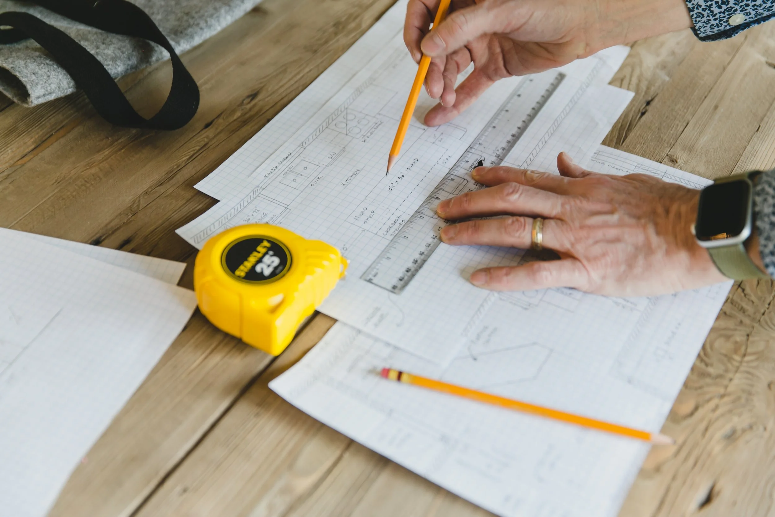 Person drawing on architectural plans with a pencil, ruler, and a yellow measuring tape on a wooden table. Papers, a pencil, and a smartwatch are also on the table.