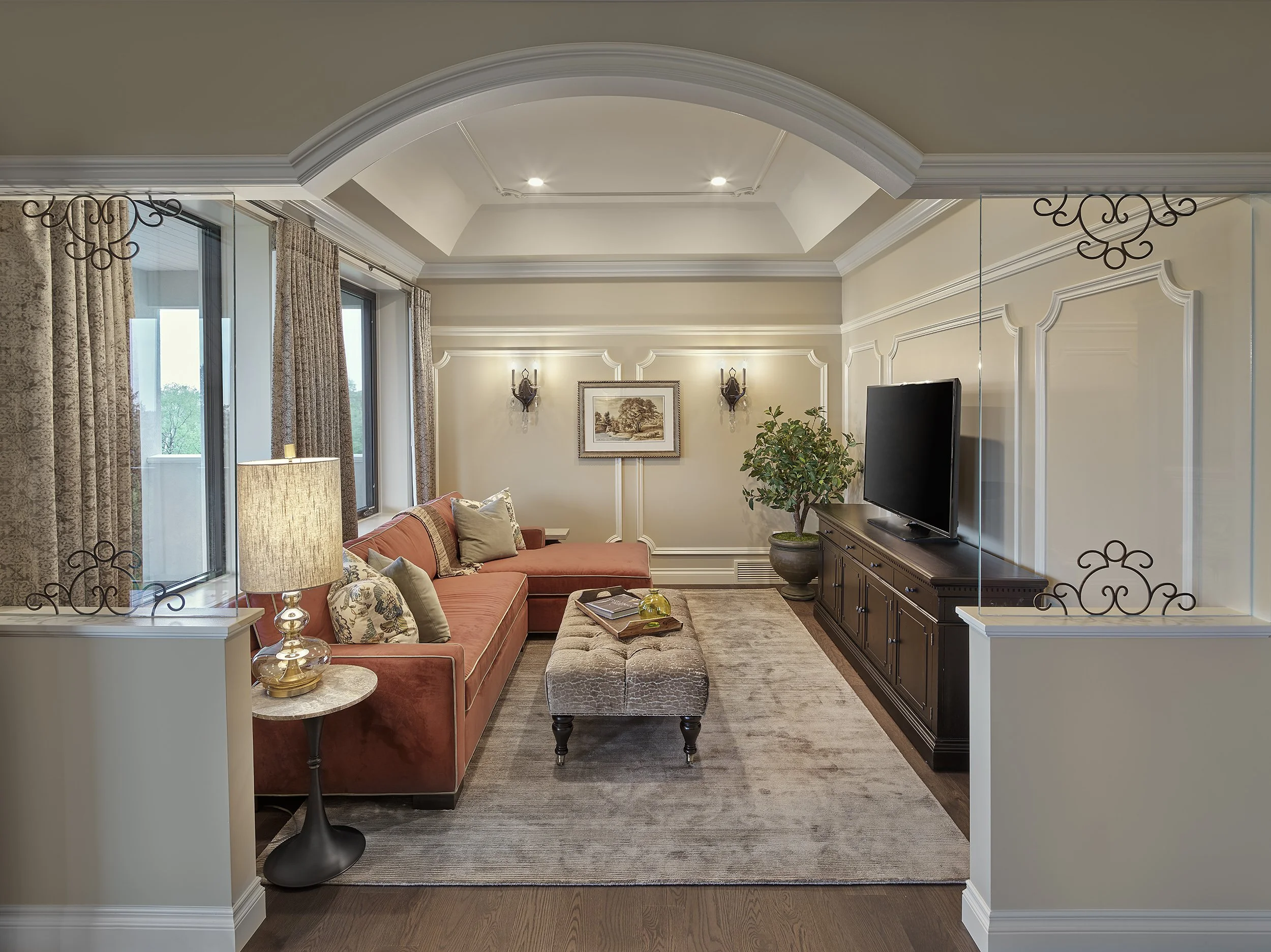 Living room with coral sectional sofa, beige ottoman, beige walls with decorative molding, dark wood TV console, flat-screen TV, window with drapes, wall sconces, framed picture, potted plant, floor lamp, area rug, hardwood floor.