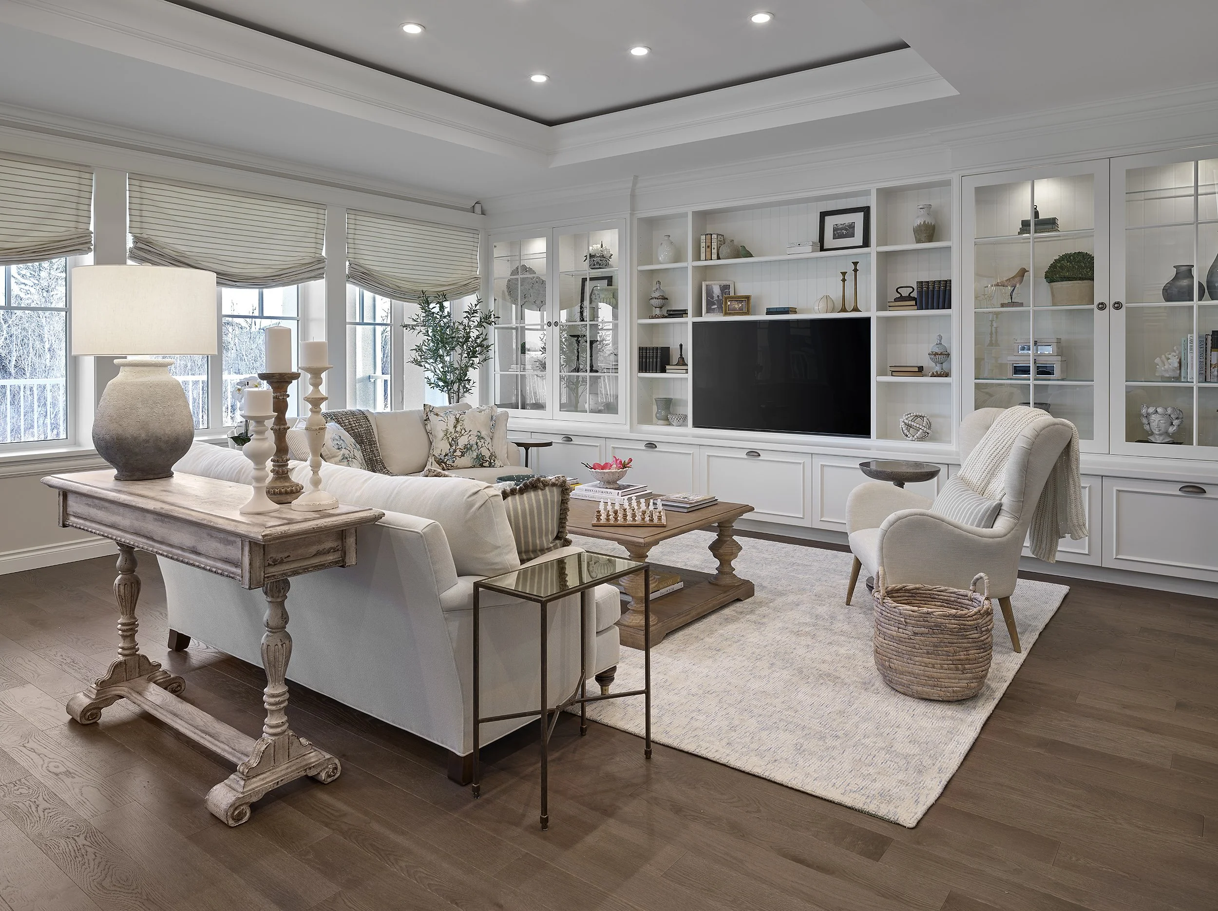 Living room with white built-in shelves, a white sofa, an armchair, wooden coffee table, and decorative items including books, vases, and picture frames. Large windows with beige Roman shades, hardwood flooring, and ceiling recessed lighting.