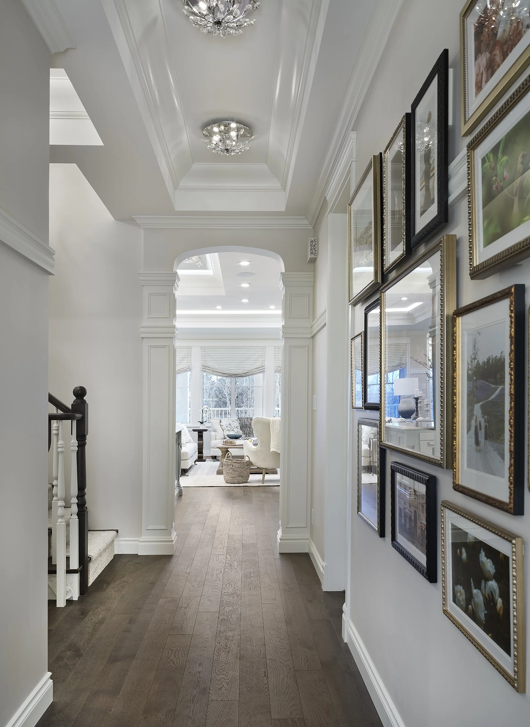 A hallway with a dark wood floor, white walls, and a gallery wall with framed photographs. A living room with large windows and white upholstered furniture is visible at the end of the hallway.