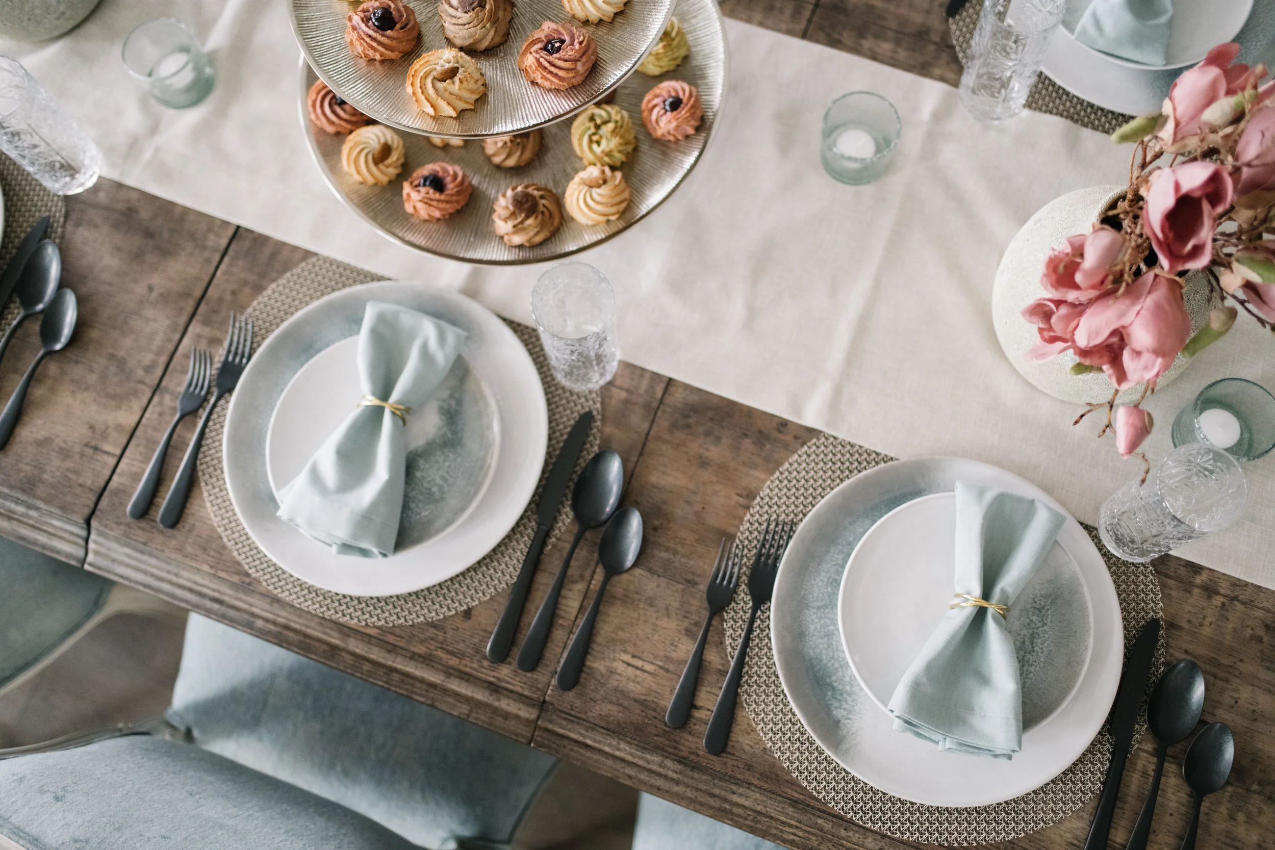 A top-down view of a dining table set for two with white plates, pale green napkins tied with gold rings, black utensils, and glassware. There is a beige table runner over a wooden table, with a silver tiered tray holding assorted cookies and pastrie
