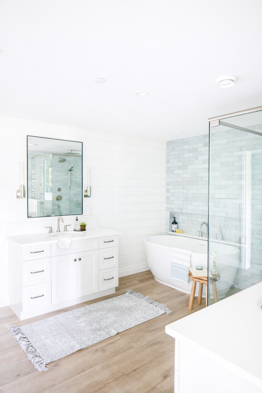 Bright, modern bathroom with white vanity, large mirror, bathtub, glass shower, and wood flooring.