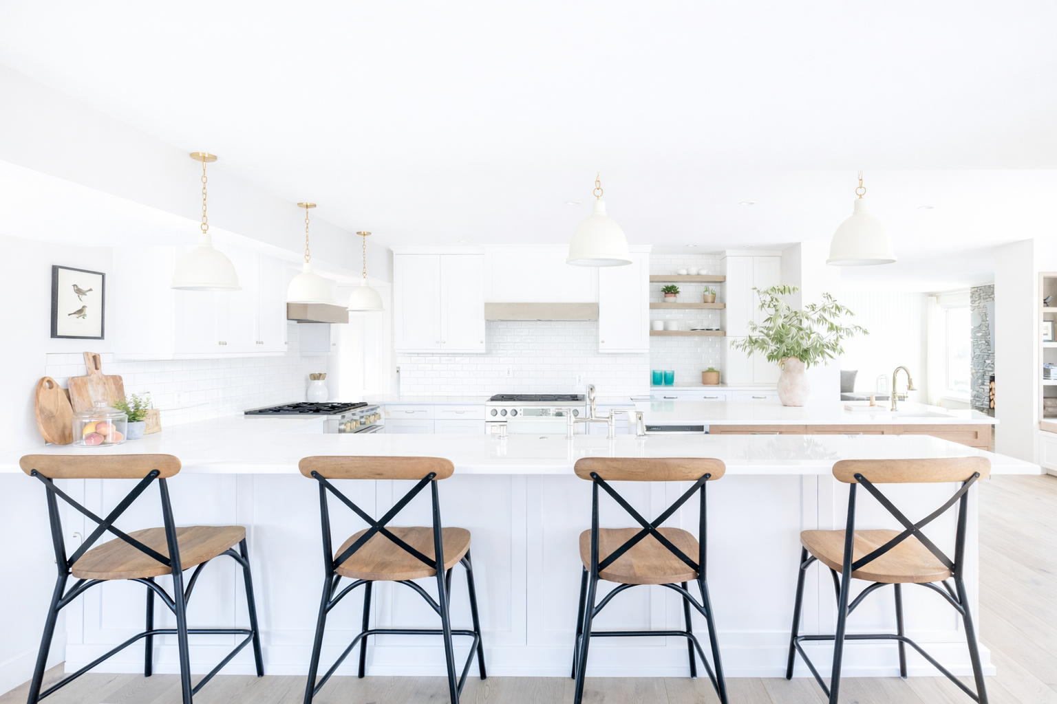 Bright, white kitchen with a large island, four wooden barstools, pendant lights, white cabinets, open shelving, and a large potted plant.