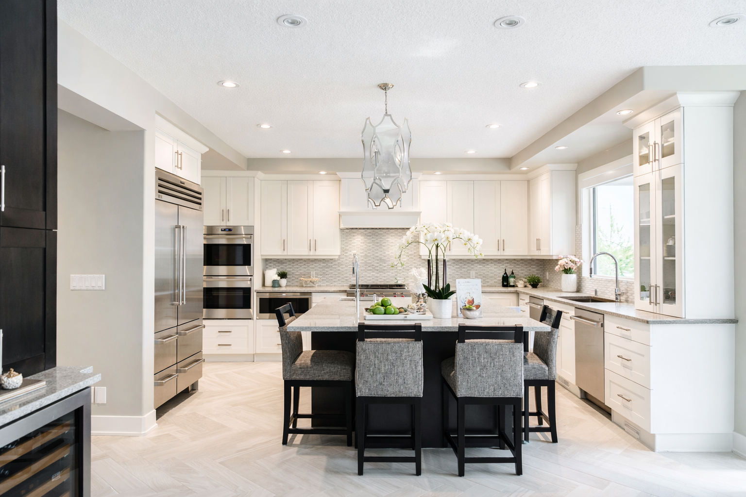 Modern kitchen with white cabinets, stainless steel appliances, black kitchen island, gray barstools, hanging light fixture, and decorative plants.
