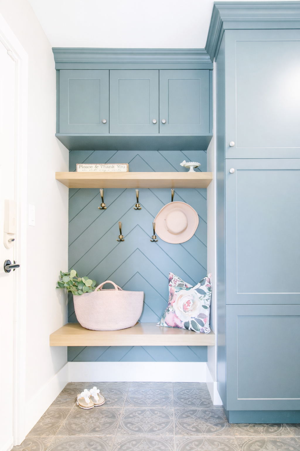 A pastel blue mudroom with wooden shelves, hooks, and cabinets, decorated with a pink hat, a floral pillow, a pottedplant, and slippers on the tiled floor.