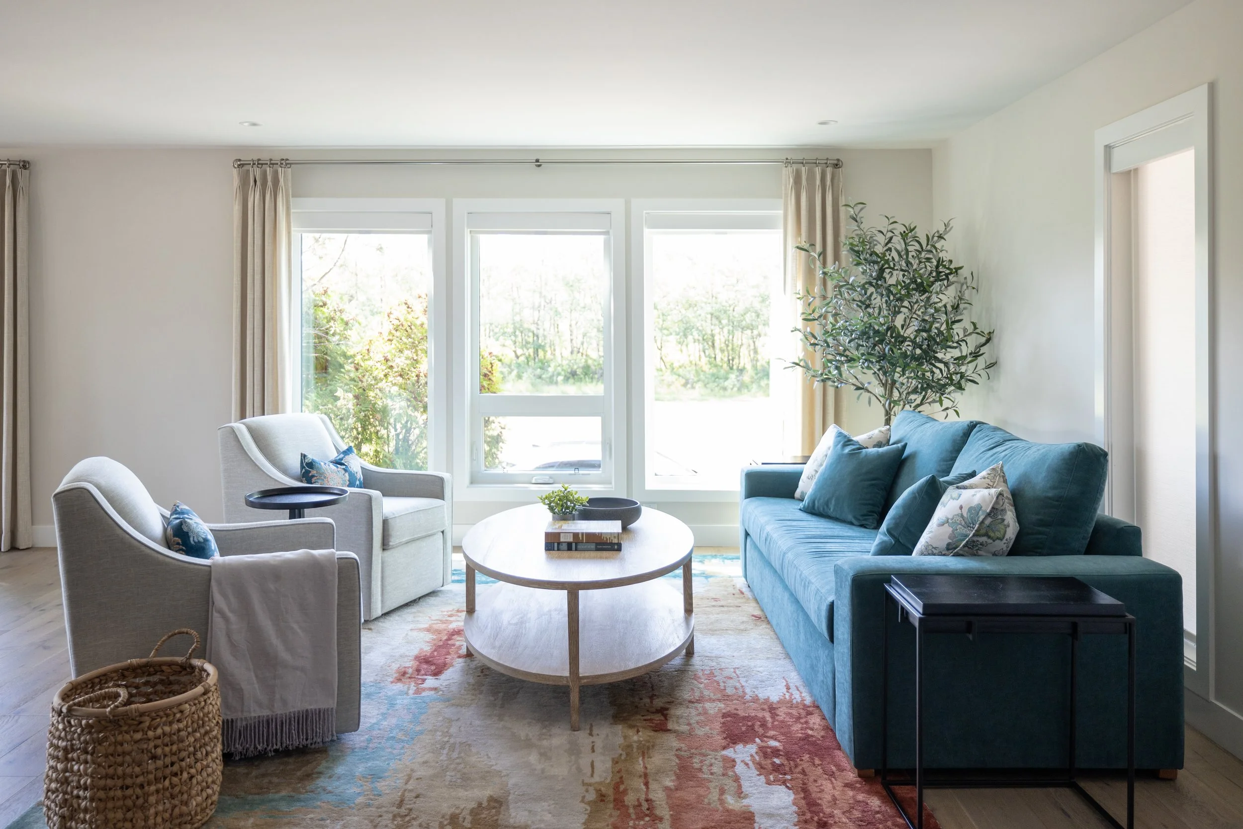 Bright living room with large windows, beige curtains, white walls, a blue sofa with cushions, two light-colored armchairs with cushions, a wooden oval coffee table with books and a plant, a small black side table, a decorative woven basket, and a large potted plant in the corner.