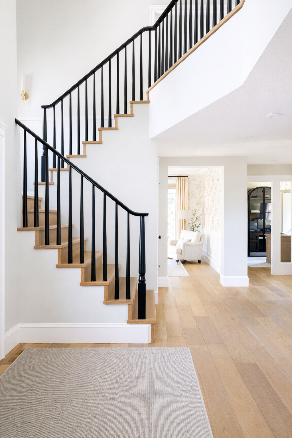 Interior of a modern home featuring a staircase with wooden steps and black metal railing, light hardwood floors, a white wall, and a living area with a white armchair and a window with curtains.