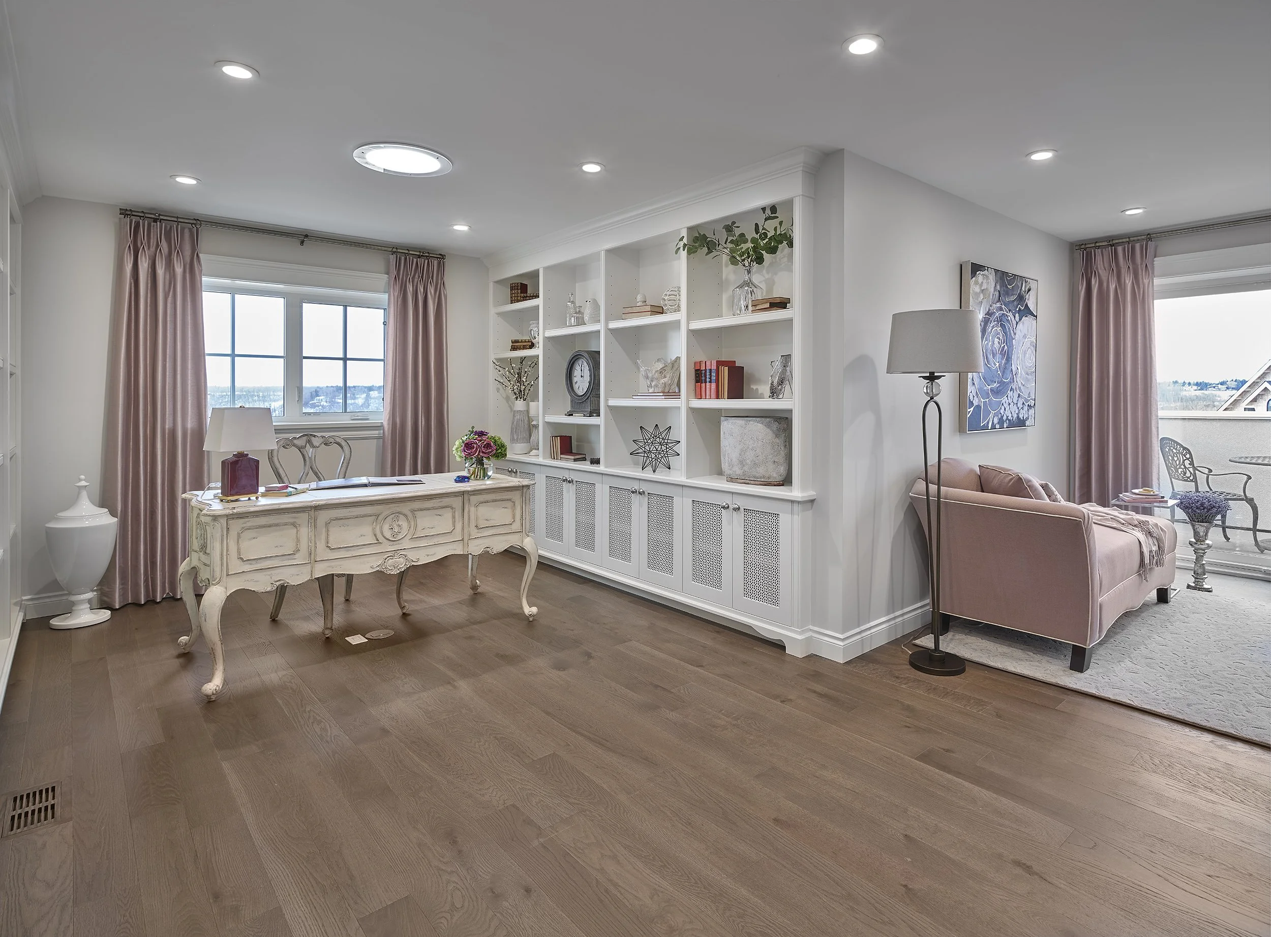 Living room with a vintage white desk, pink curtains, a built-in white bookshelf with decor, a pink sofa, a floor lamp, and a balcony with outdoor seating.