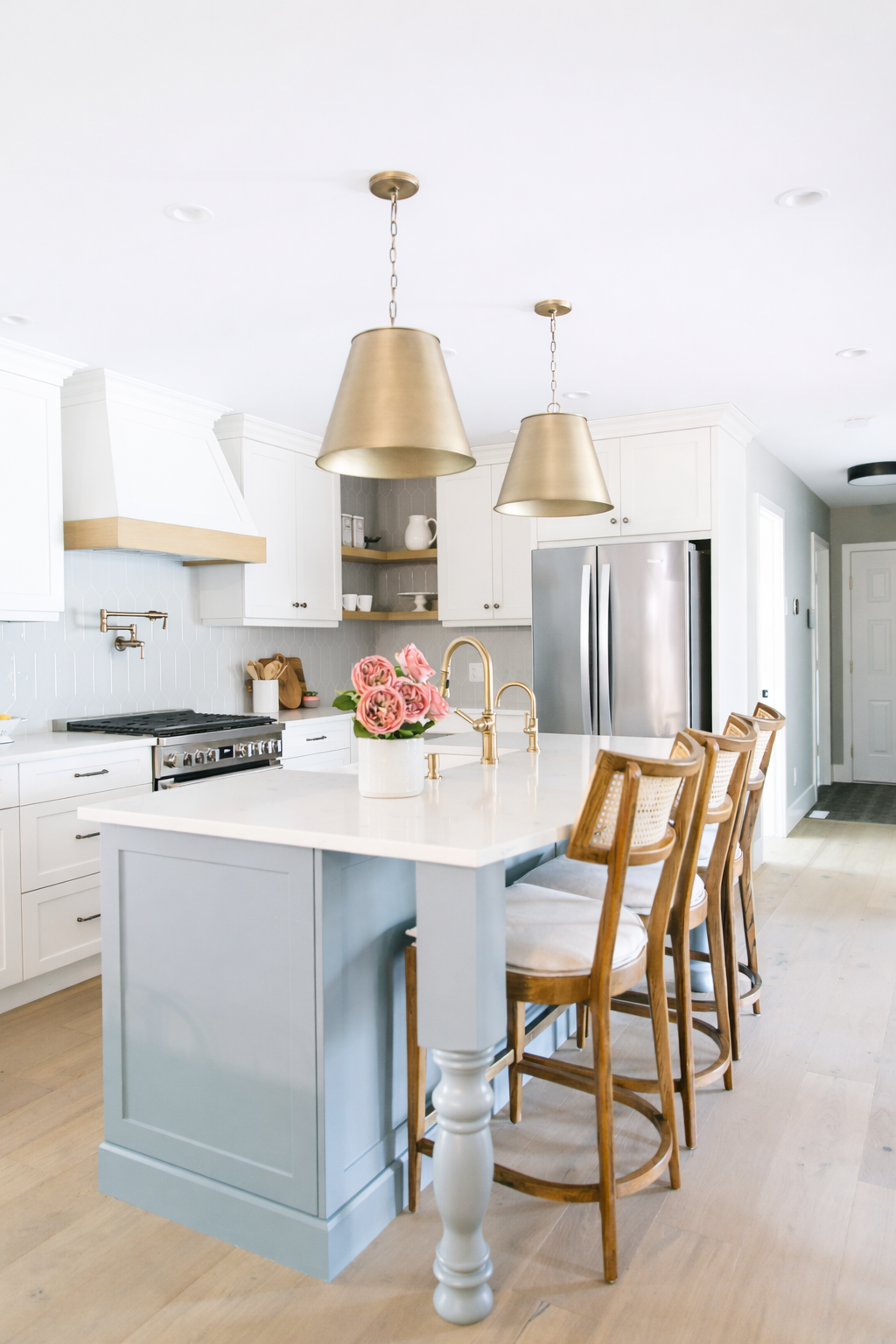 A modern kitchen with white cabinets, a kitchen island with a white marble top, and wooden chairs with wicker backs. There are brass pendant lights hanging from the ceiling, a bouquet of pink roses on the island, and stainless steel appliances, inclu