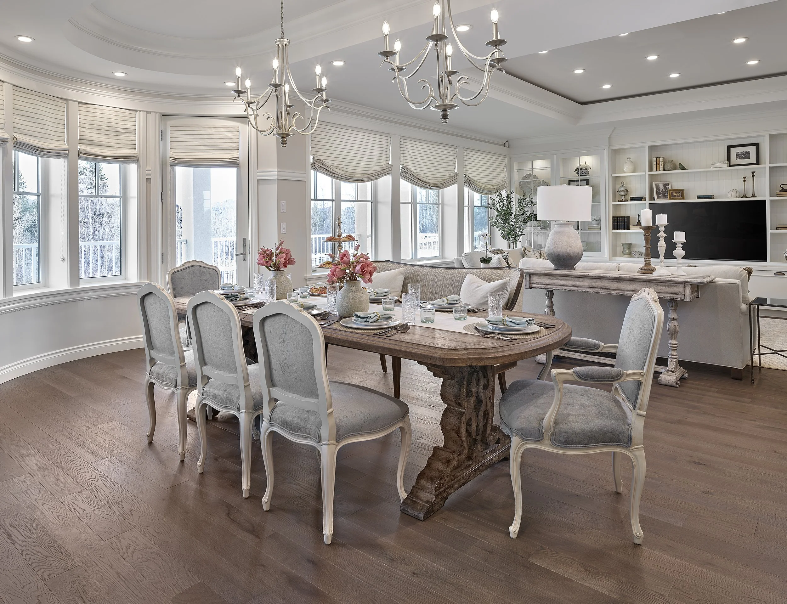 Elegant dining room with a wooden table set for a meal, surrounded by upholstered chairs, and a living area with a large white sofa, a built-in white bookshelf, and decorative accessories. Large windows with beige shades let in natural light. A chand