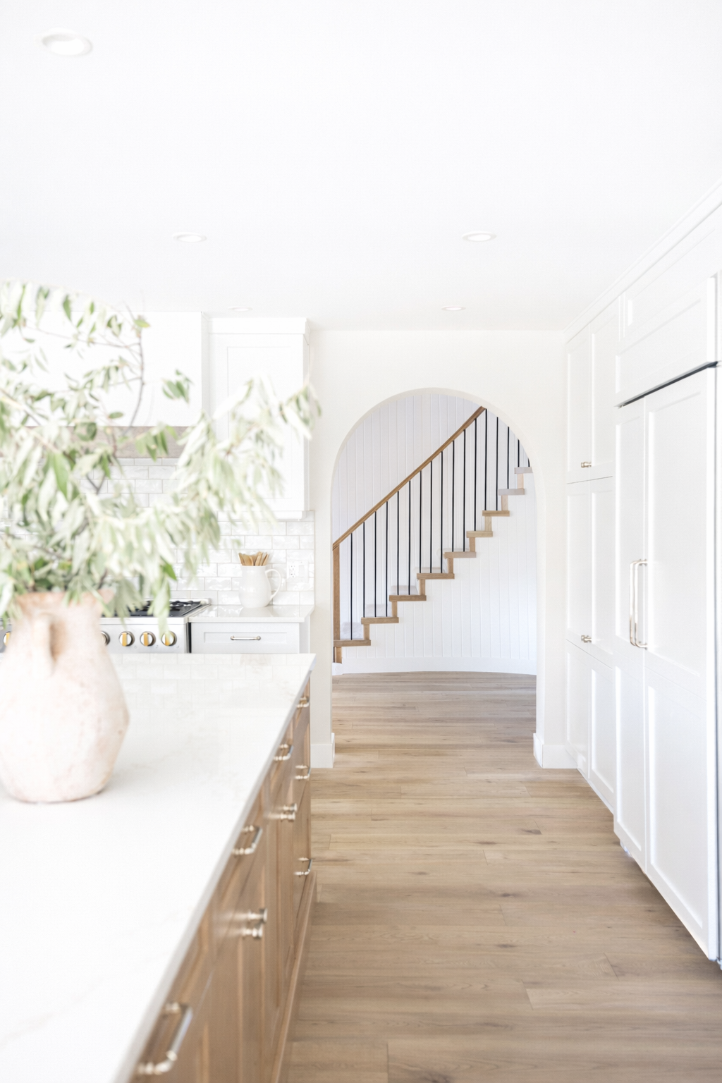 Bright modern kitchen with wooden cabinets, white countertop, large potted plant, and a view of a staircase through an arched doorway.