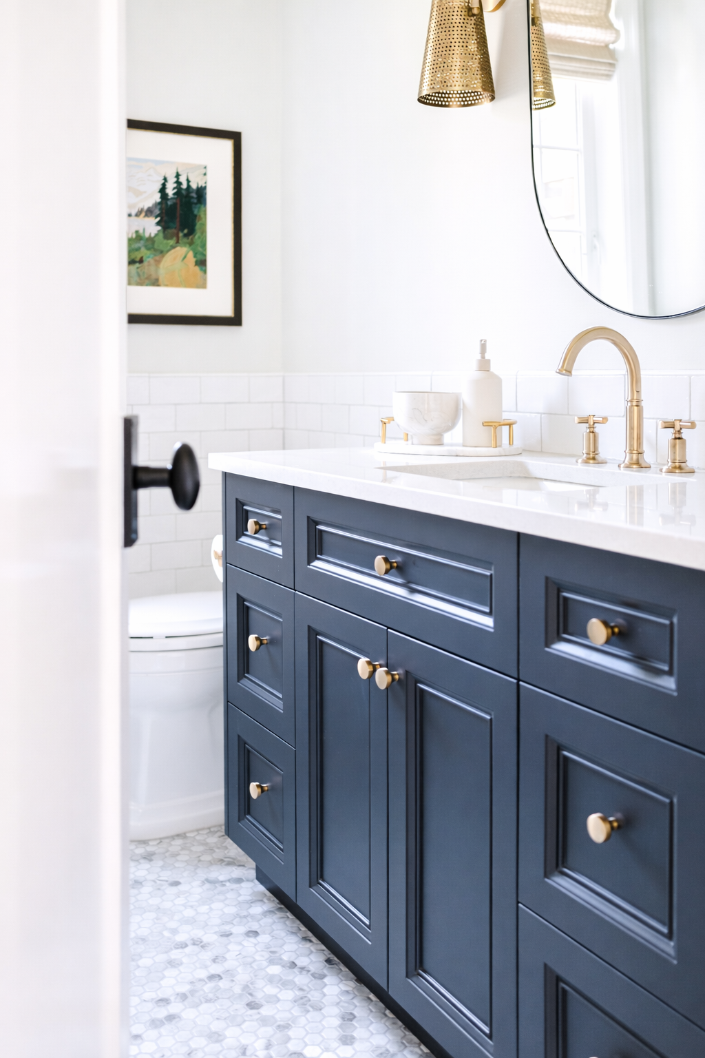 Bathroom vanity with blue cabinets, white countertop, gold hardware, and a round mirror. There are soap dispensers and a vessel sink with a gold faucet. A window and wall art are visible.