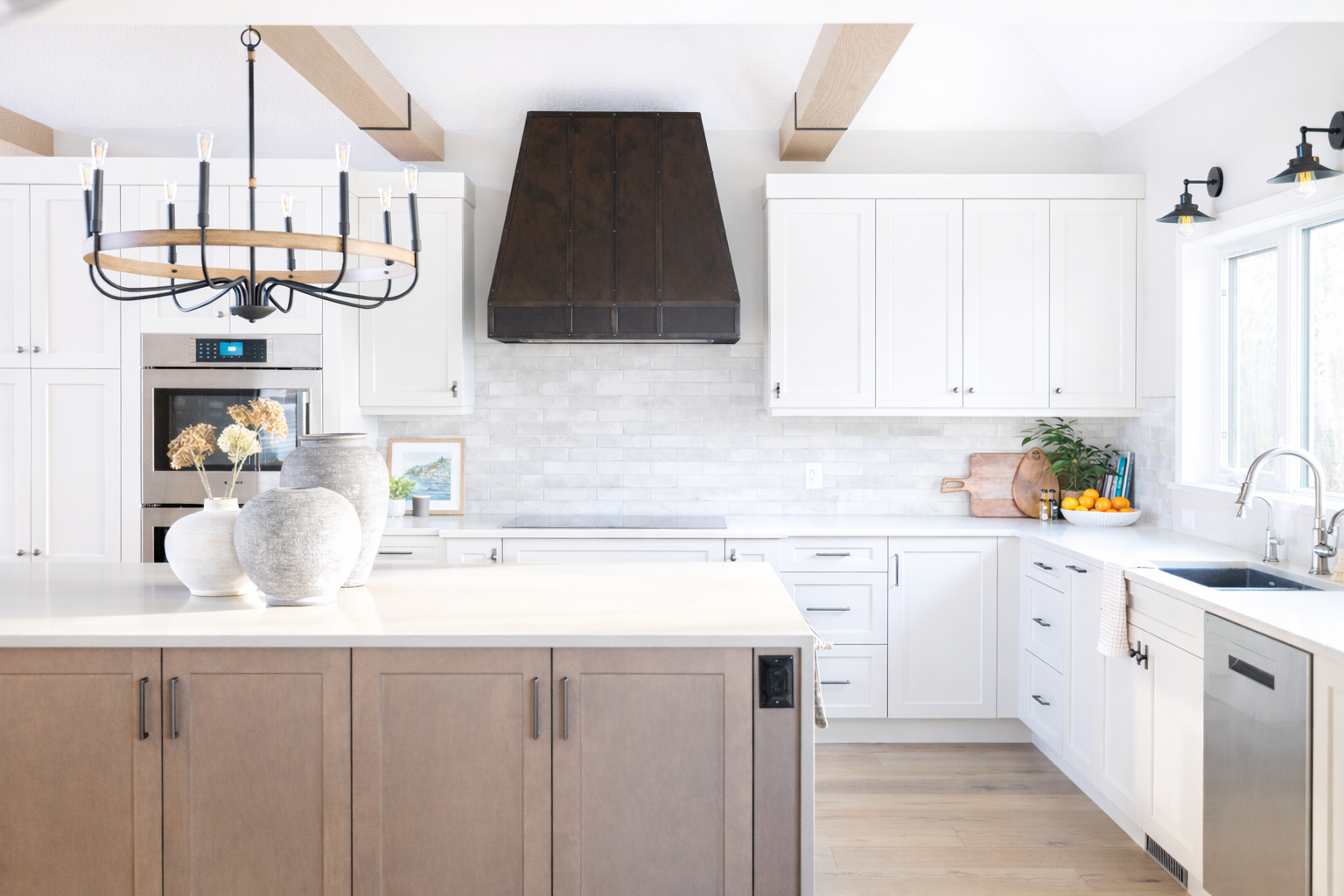 Bright kitchen with white cabinets, a central island with a beige base, and a white countertop. Features include a black range hood, a stainless steel microwave, a large window above the sink, black wall-mounted light fixtures, a black chandelier, an