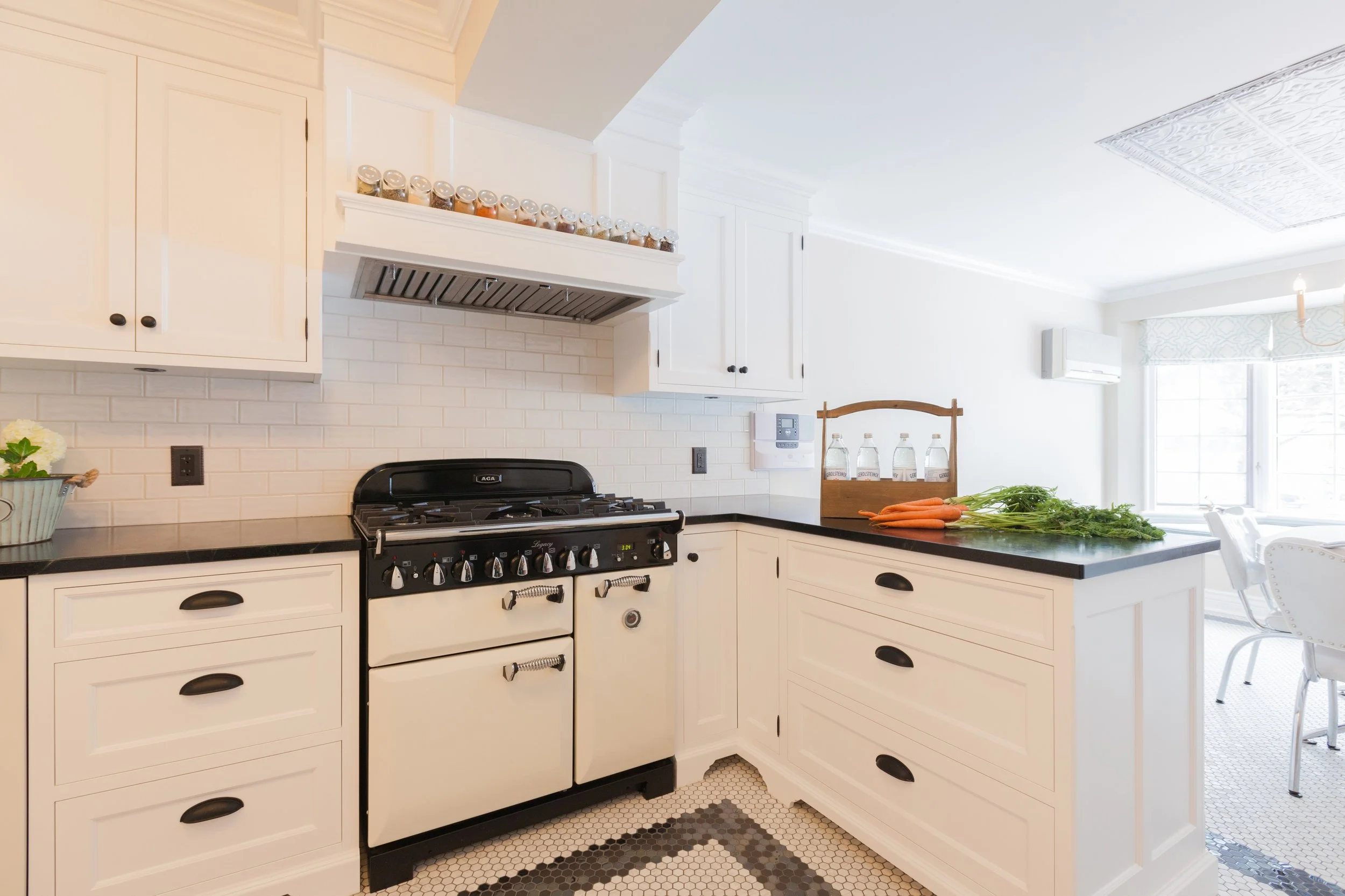 Kitchen with white cabinets, black countertops, a black and cream stove, a spice jar shelf above the stove, and a cutting board with carrots and greens on the counter.