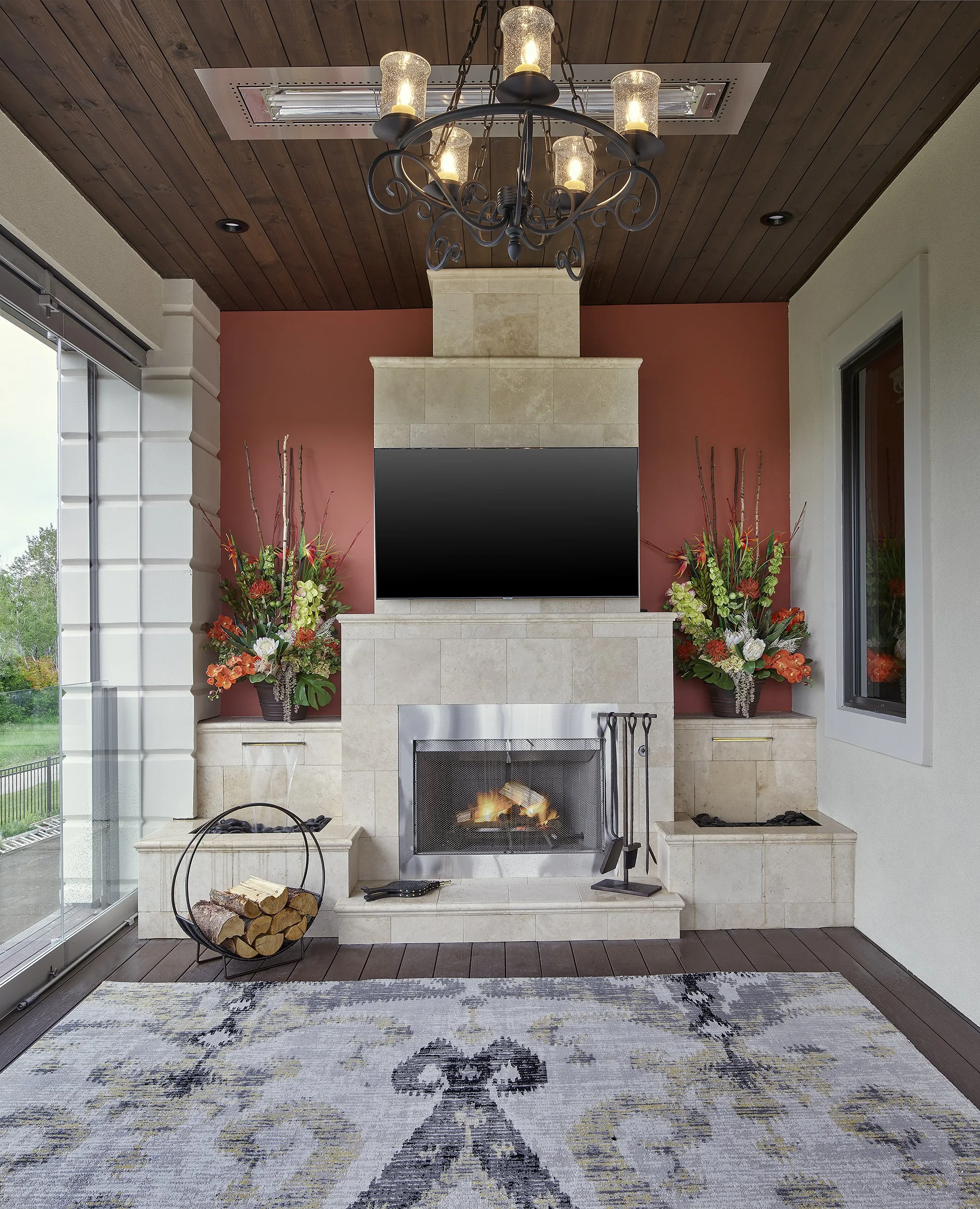 Living room with beige stone fireplace, mounted flat-screen TV, floral arrangements, logs, and fireplace tools, with dark wood ceiling and chandelier, and large glass sliding door.