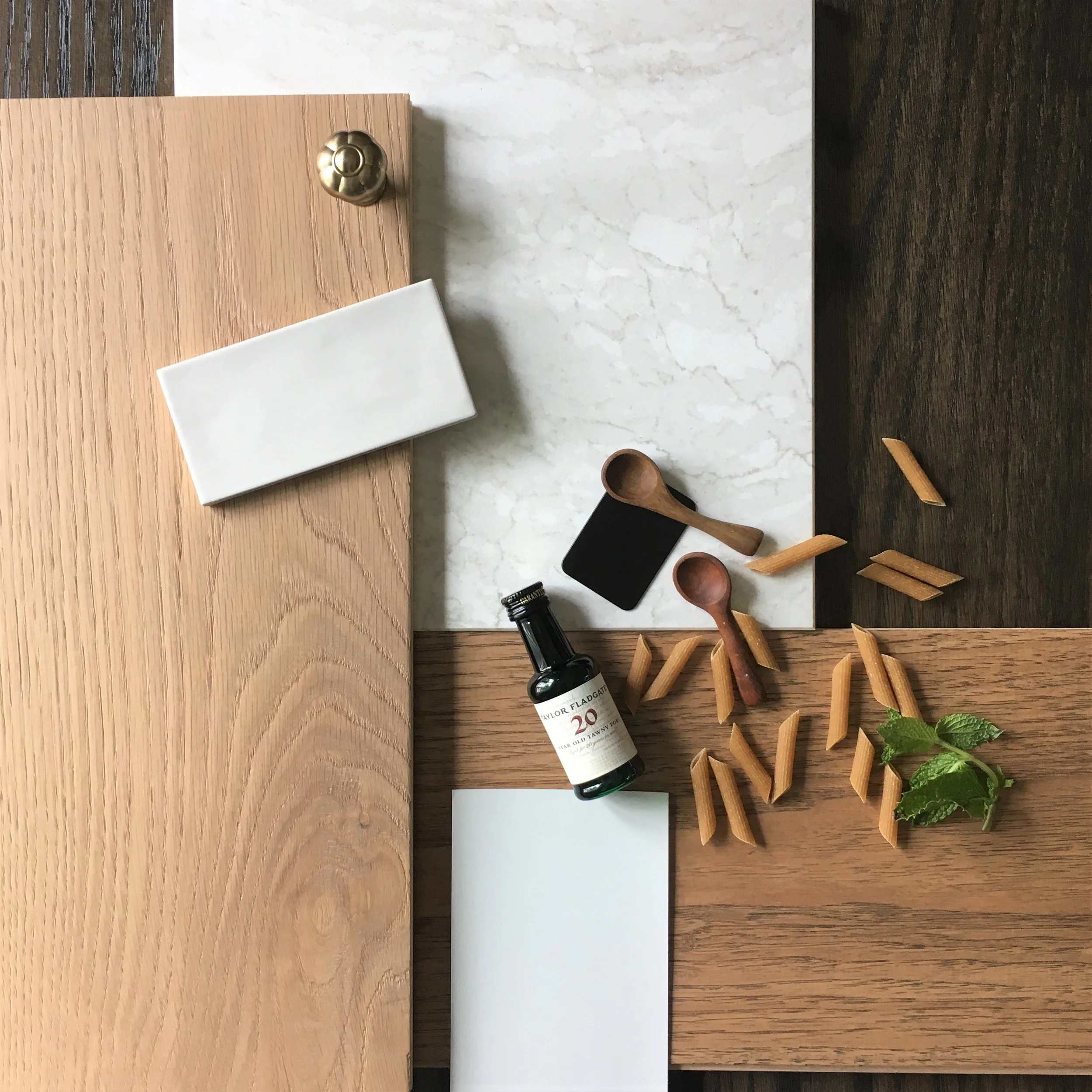 A wooden table with scattered small wooden spoons, a small black cutting board, a bottle of Taylor Fladgate port wine, a white notepad, a small white box, and some green mint leaves.