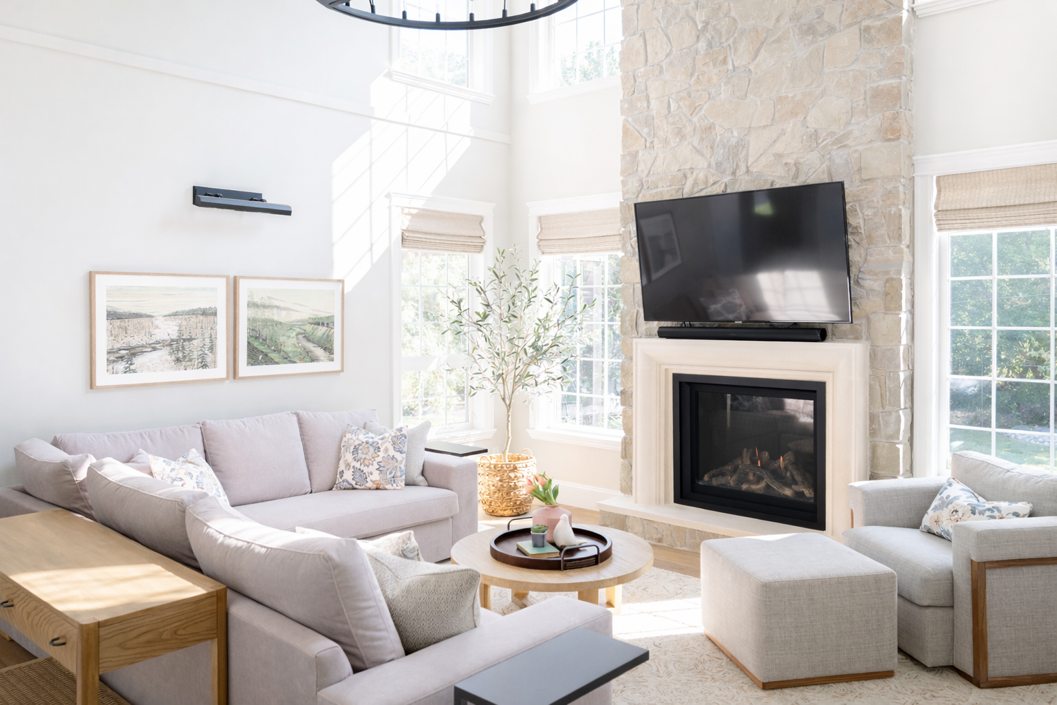 Bright living room with large windows, a stone fireplace with a TV mounted above, a light-colored sofa, an ottoman, a round coffee table with decorative items, and a potted plant near the windows.