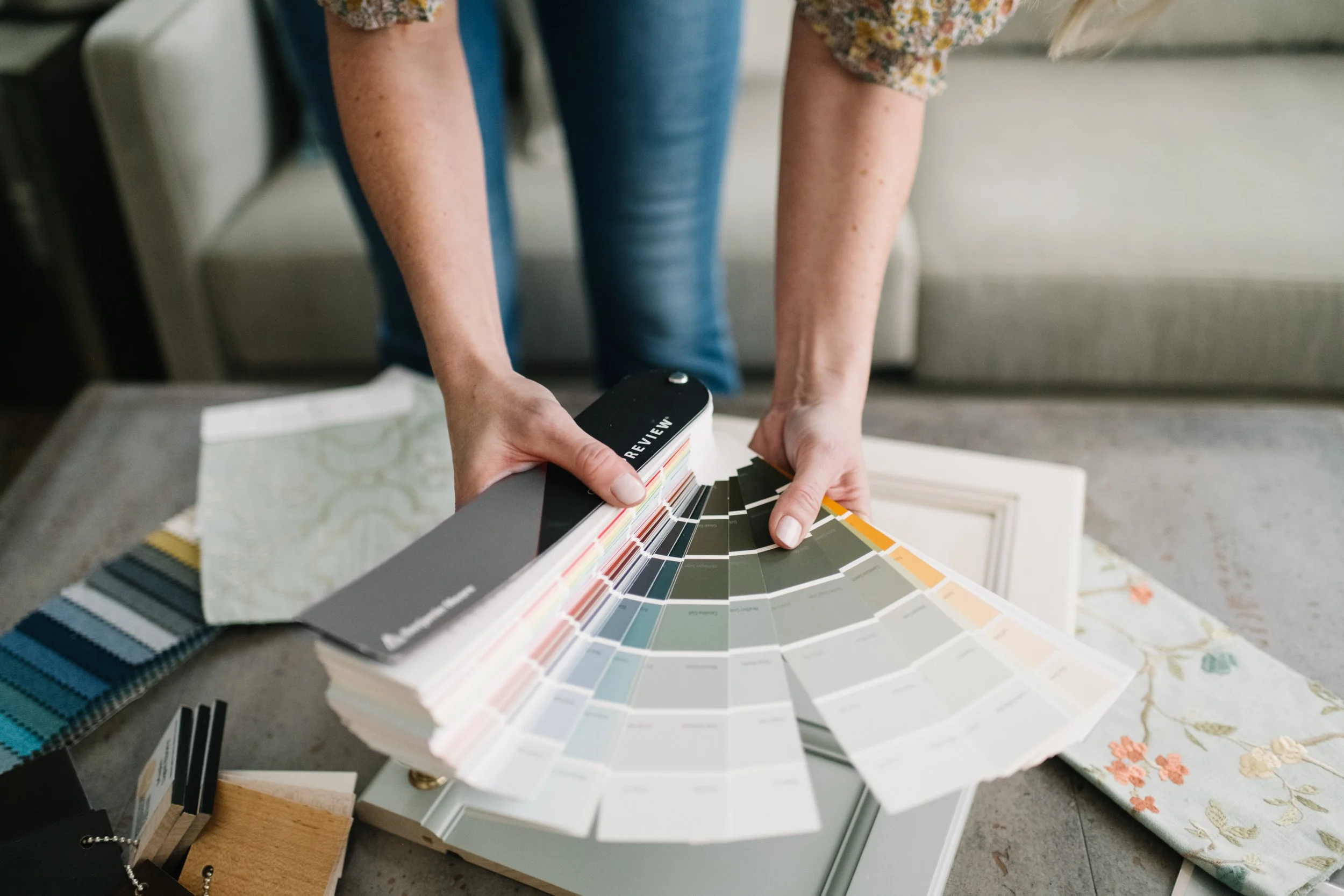 Person holding a fan deck of paint color samples over a table with fabric swatches and a framed picture.