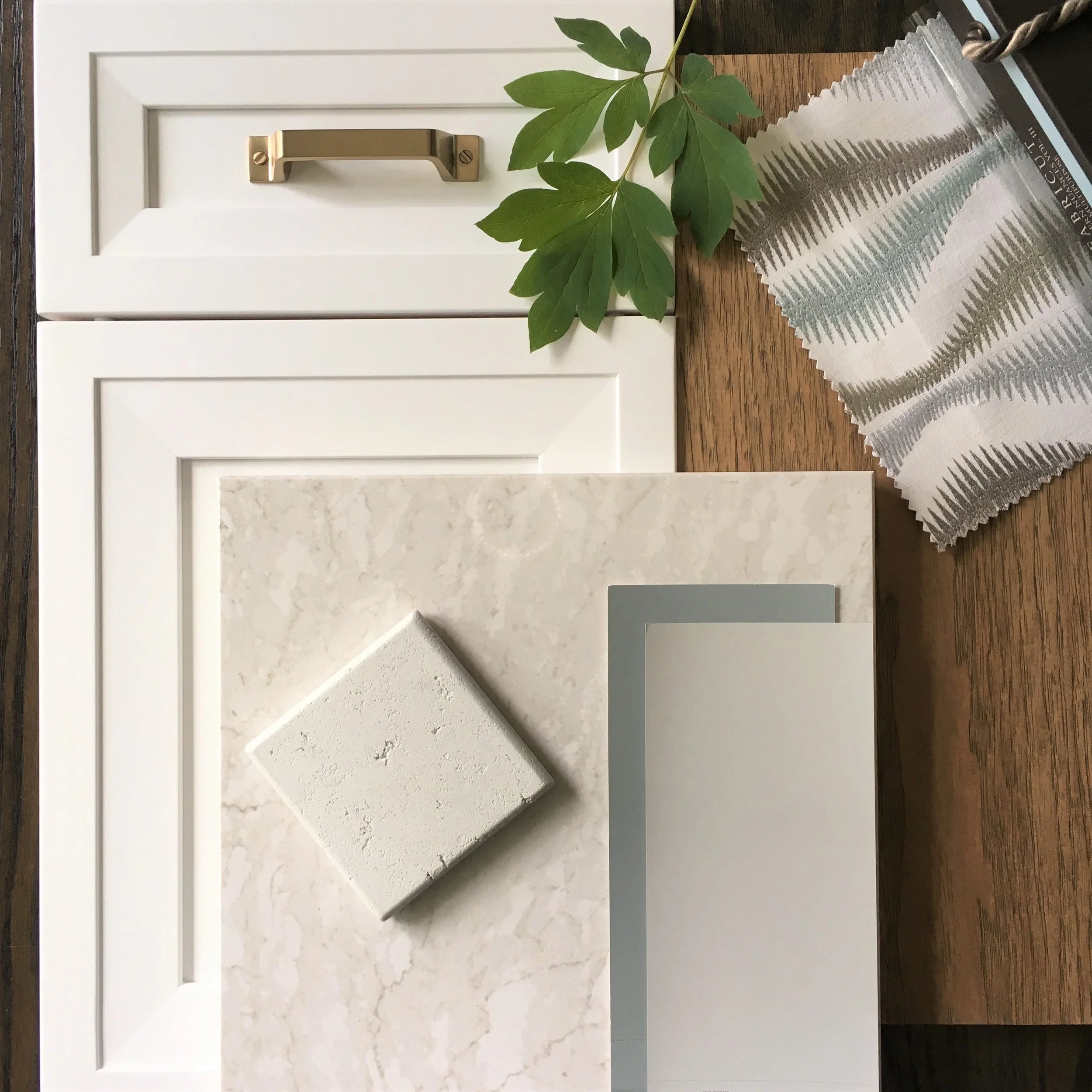 A collection of neutral-colored interior design materials including a beige marble tile, a small textured white tile, a light gray paint sample, and a white cabinet door with a brass handle. There is a green leafy branch, a fabric sample with green and beige fern pattern, and a wooden surface in the background.