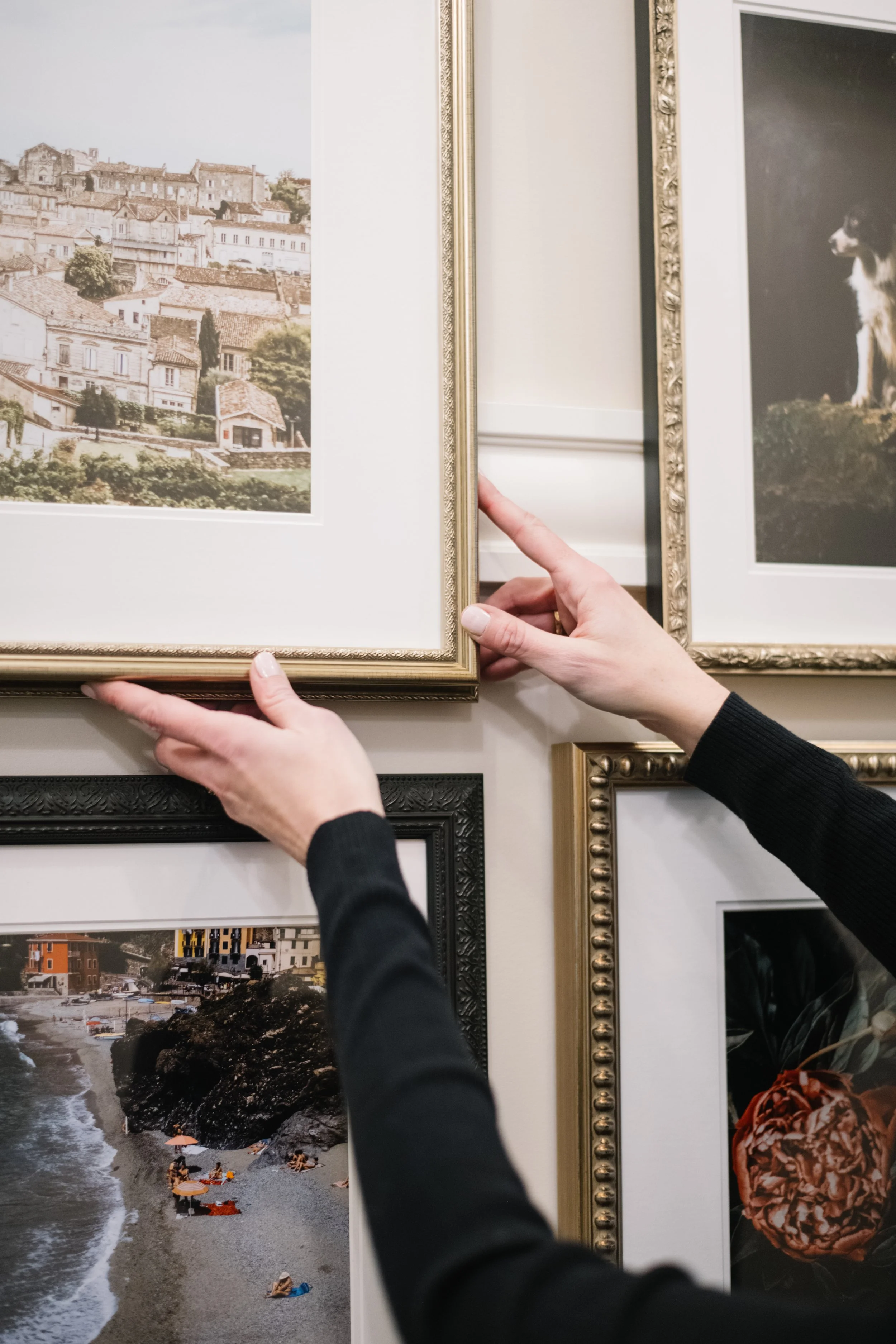 A person hanging a framed picture of a town on a wall decorated with other framed photographs.