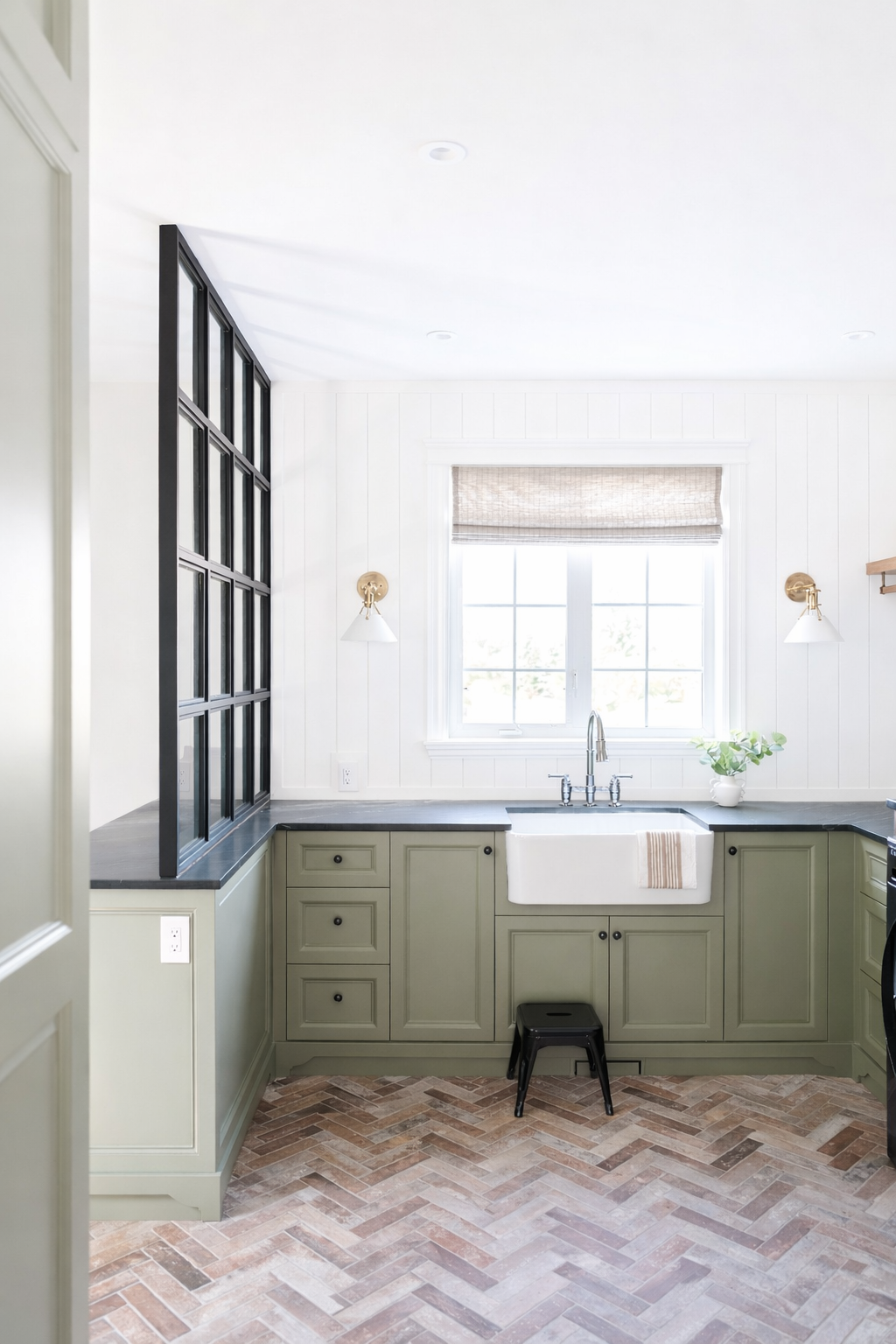 Bright kitchen with white walls, black countertops, green cabinetry, and a large window above the sink, with a small black stool in front of the cabinets.