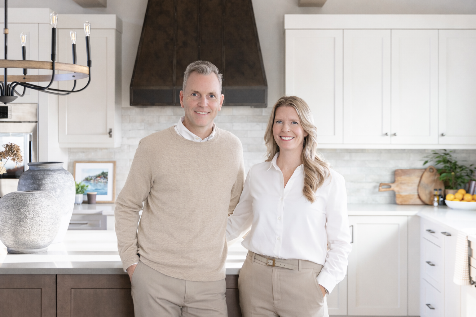 A smiling man and woman stand in a modern kitchen. The man is wearing a beige sweater over a white collared shirt, and the woman is dressed in a white blouse with beige pants. They are standing close together, smiling at the camera.