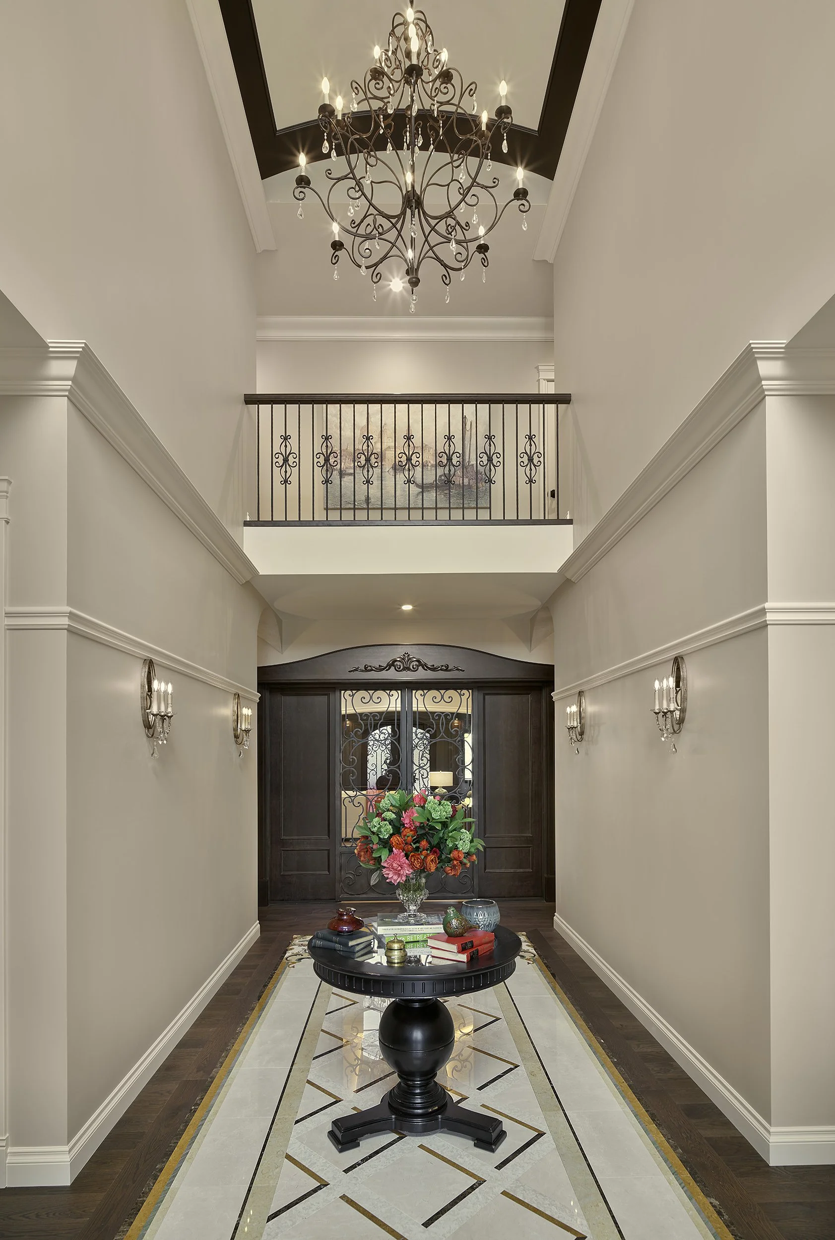 Elegant foyer with chandelier, decorative wall sconces, a black pedestal table with a bouquet of colorful flowers, and a dark wooden front door with ornate ironwork.