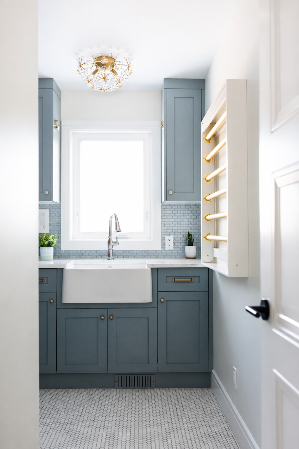 Modern kitchen with blue cabinets, a white farmhouse sink, gray backsplash, and a window above the sink. There are two potted plants on the counter and a white wall with a gold hooks rack.