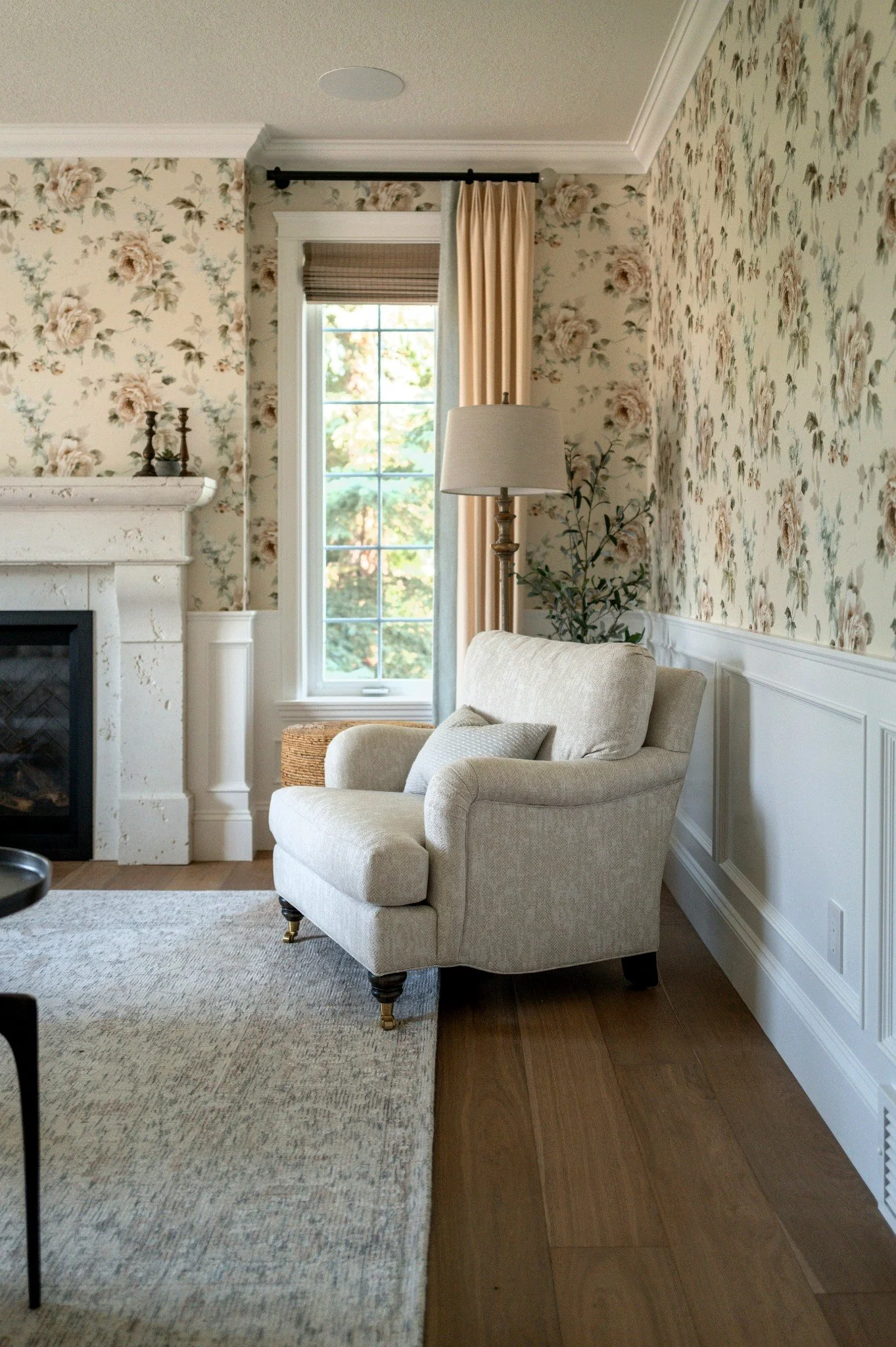 Living room corner with a beige armchair with a cushion, a floor lamp, a window with a Roman shade and curtains, a floral wallpapered wall, a gina fireplace, and a potted plant.