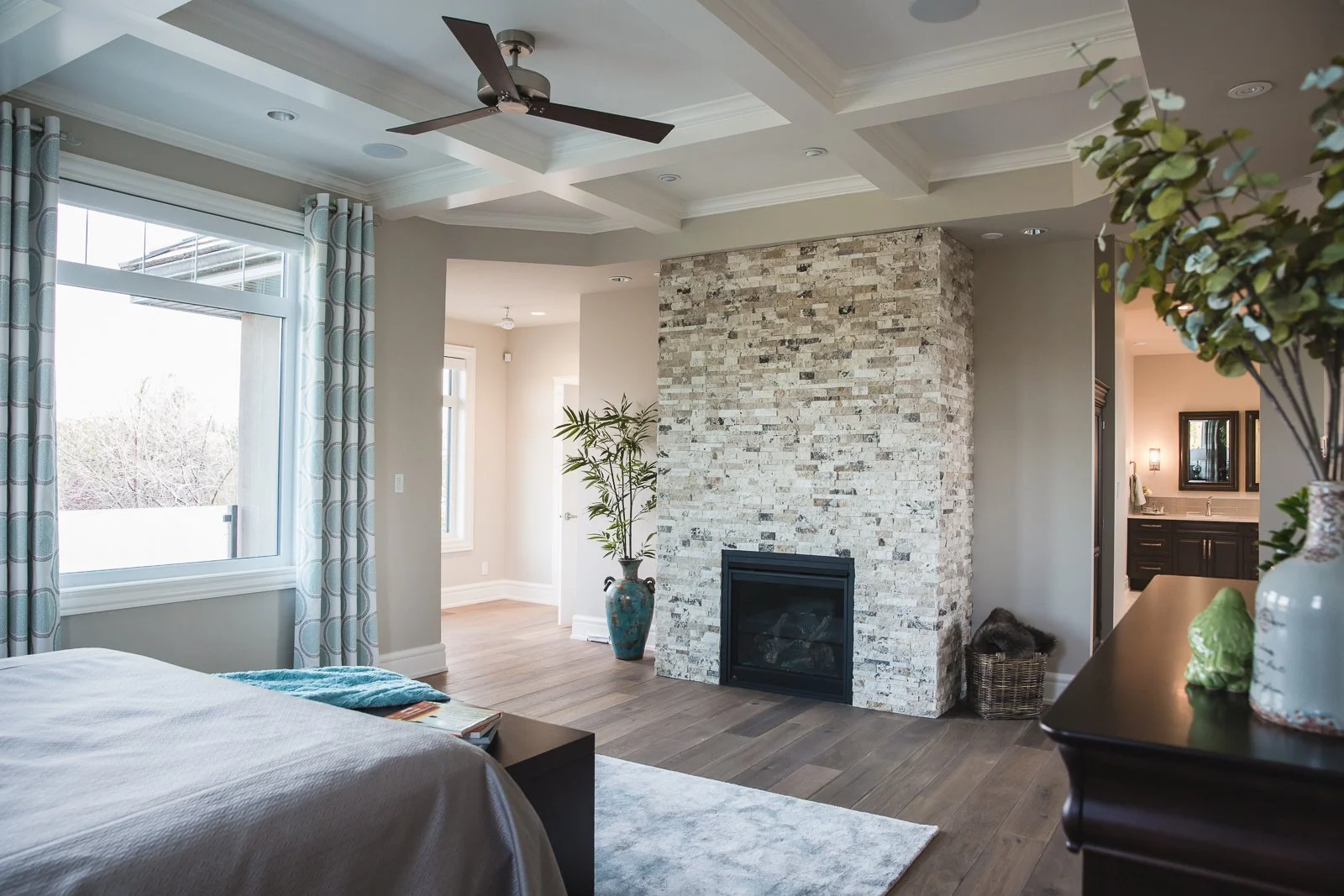 Living room with a stone fireplace, large windows with patterned curtains, ceiling fan, and hardwood floors. There are decorative vases and plants, and a glimpse of a bathroom with dark cabinetry in the background.
