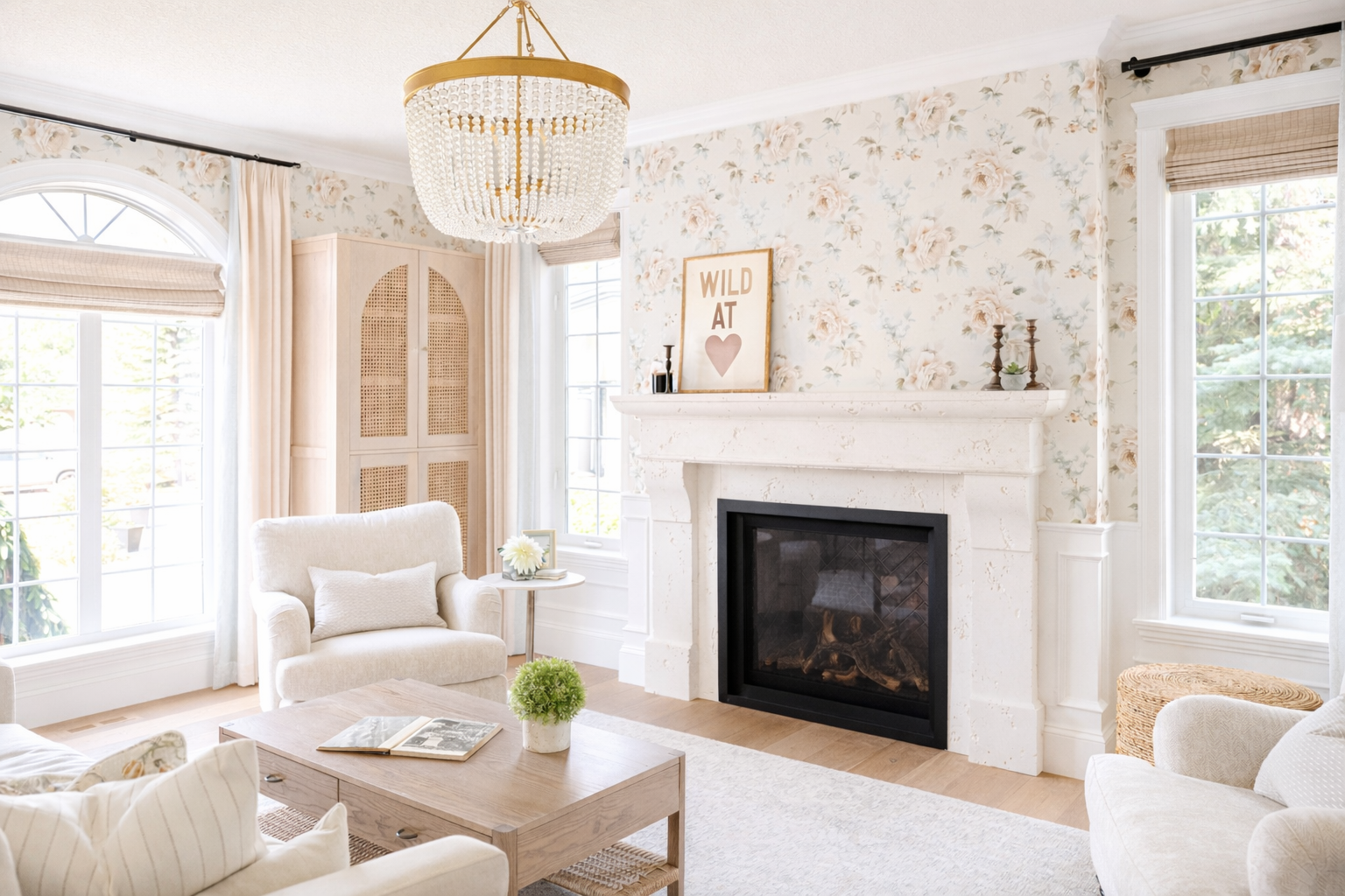Bright living room with floral wallpaper, large windows, a white armchair, a fireplace, and a chandelier.