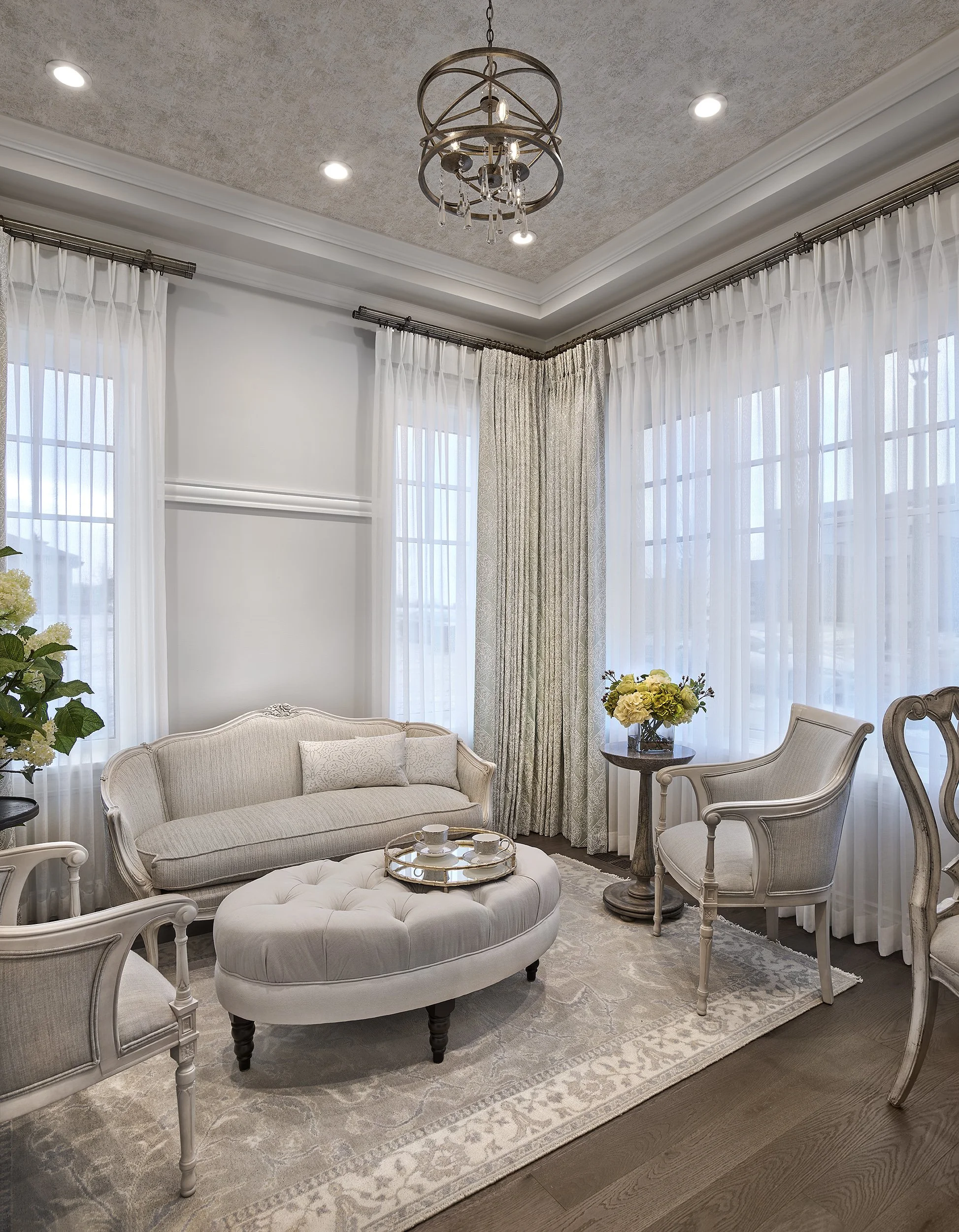 Elegant living room with large windows, white curtains, a beige upholstered sofa, a tufted ottoman with tea cups, a side table with yellow flowers, and vintage-style chairs.
