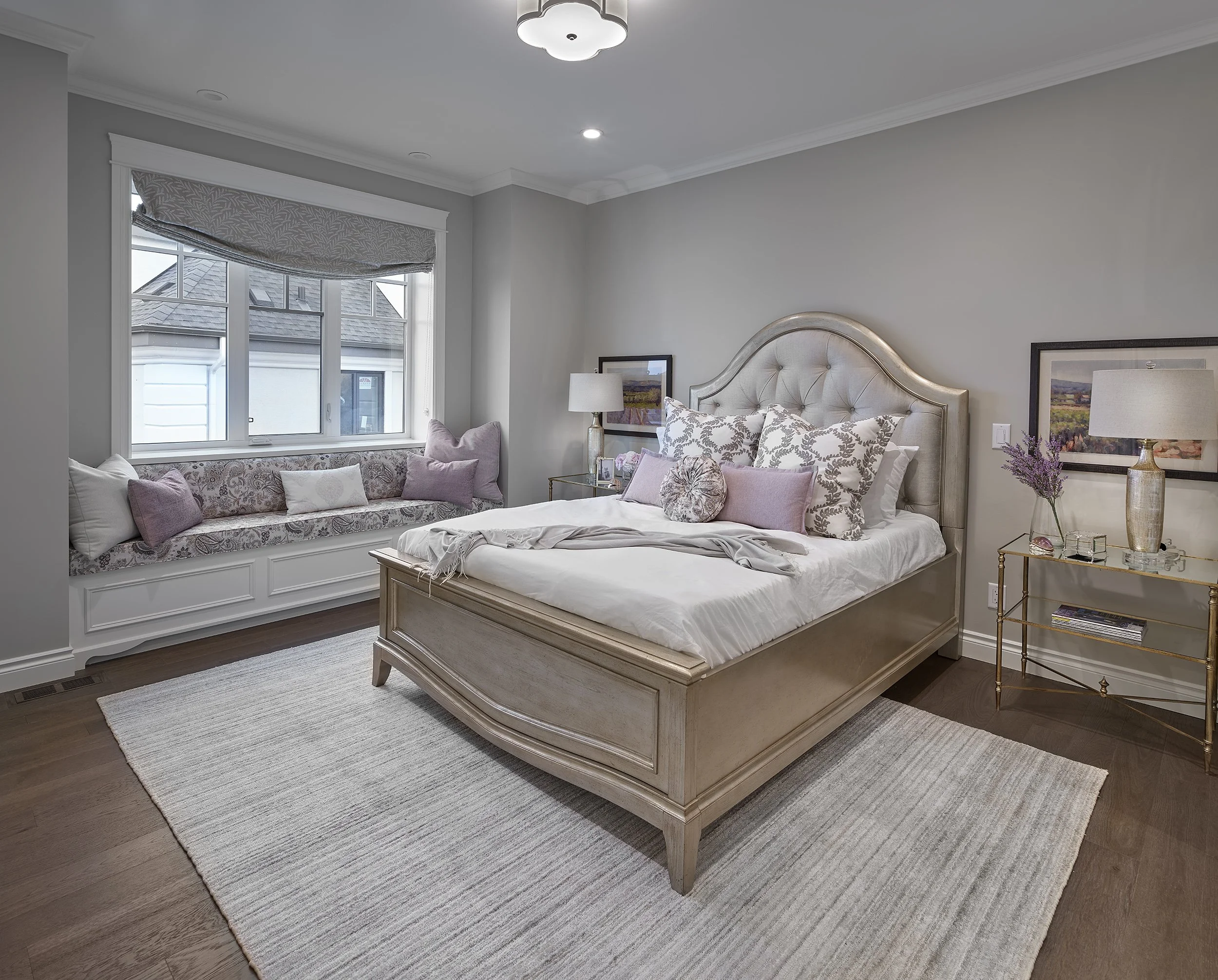A bedroom with a large bed featuring an ornate headboard, decorated with pink and floral patterned pillows, and a window seat with cushions underneath a window. There are two matching bedside lamps and framed landscape artworks on the walls.