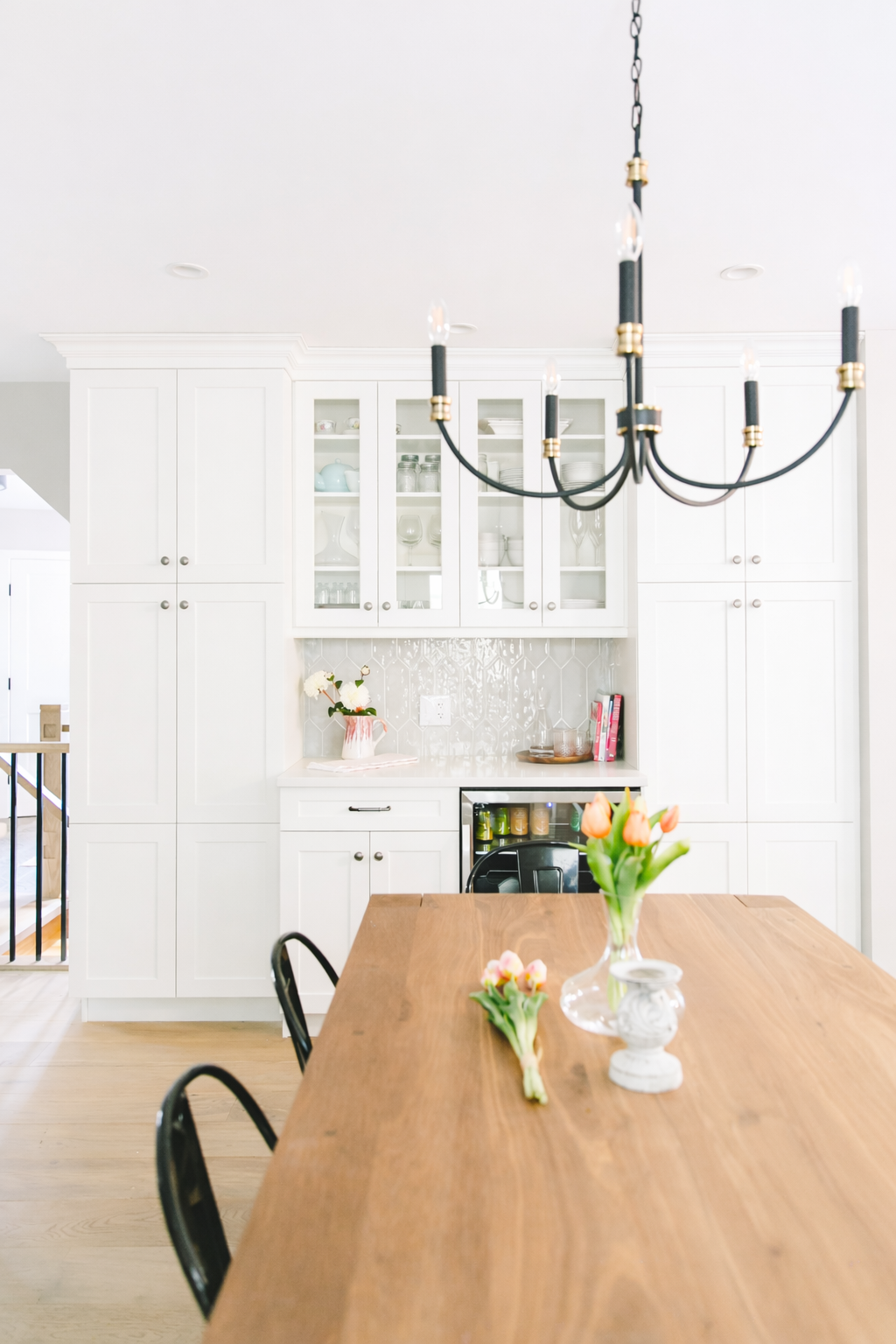 Bright kitchen with white cabinetry, a wooden dining table with flowers, and a black chandelier.