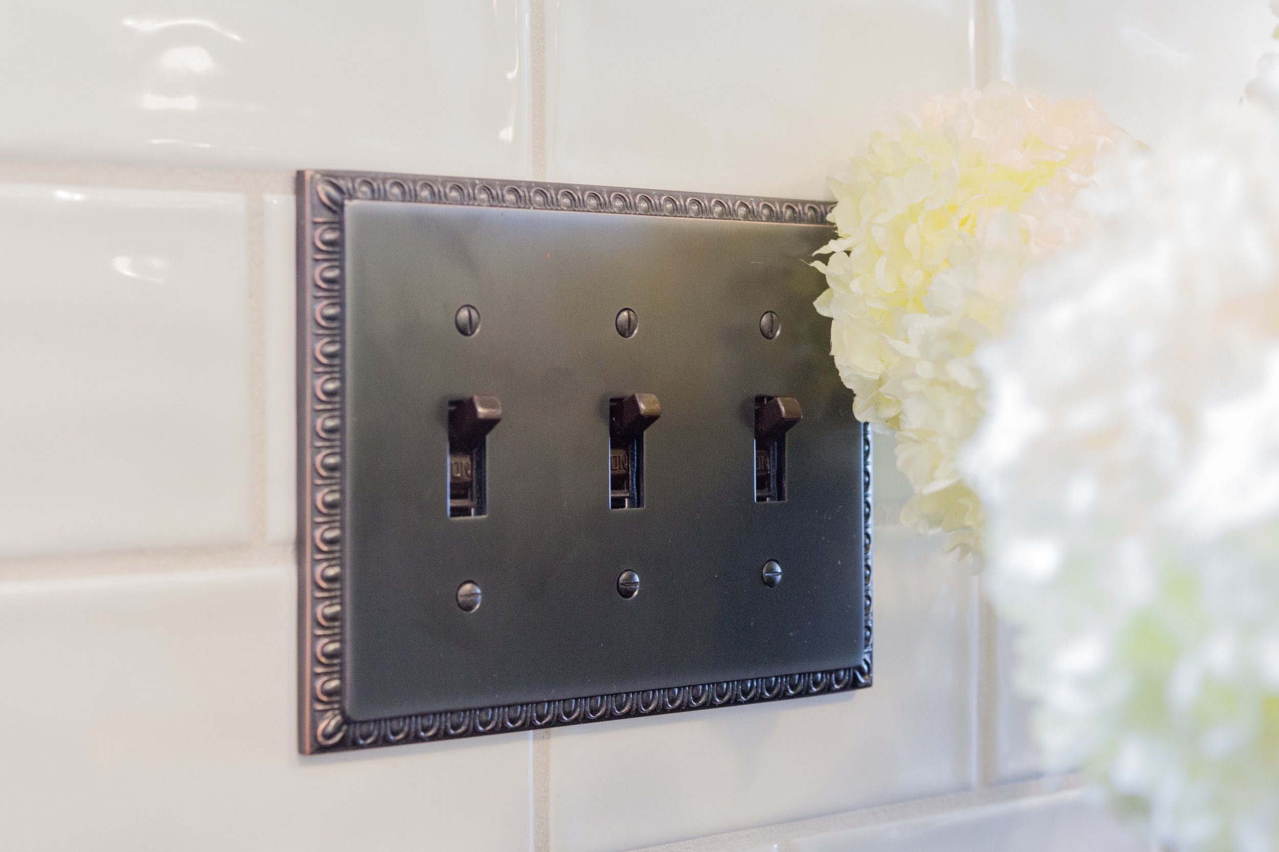 Black decorative triple light switch on a white tiled wall with white flowers to the right.