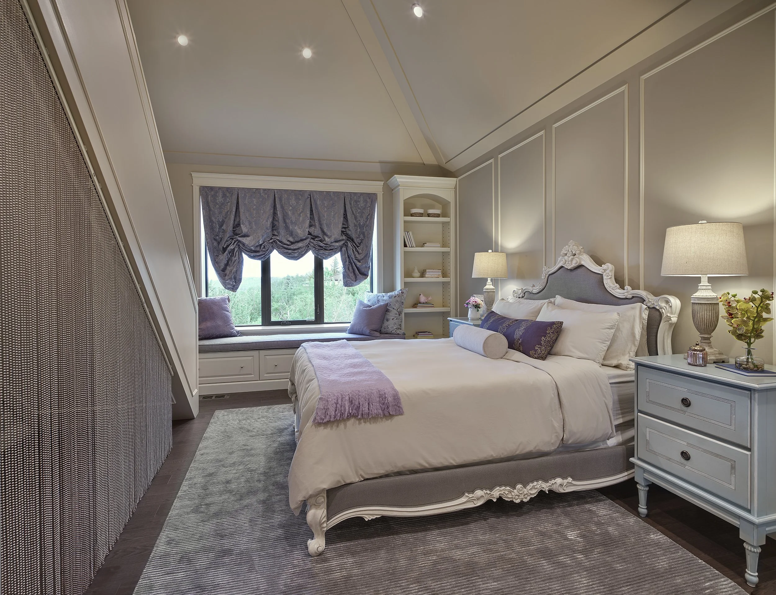 A cozy bedroom with a large bed featuring a tufted headboard, nightstand with a lamp, and a window seat with pillows and a window view of greenery.