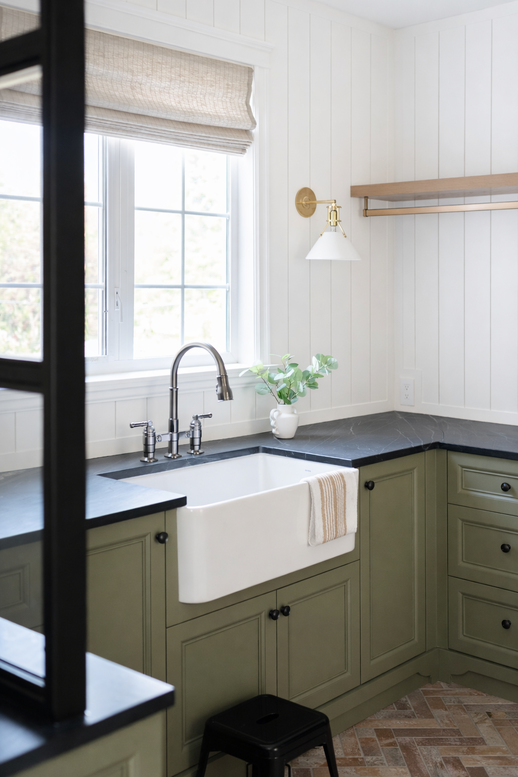 Kitchen with green cabinets, black countertop, farmhouse sink, window with beige Roman shade, small plant in a white vase, wall-mounted light fixture, and wooden shelf.