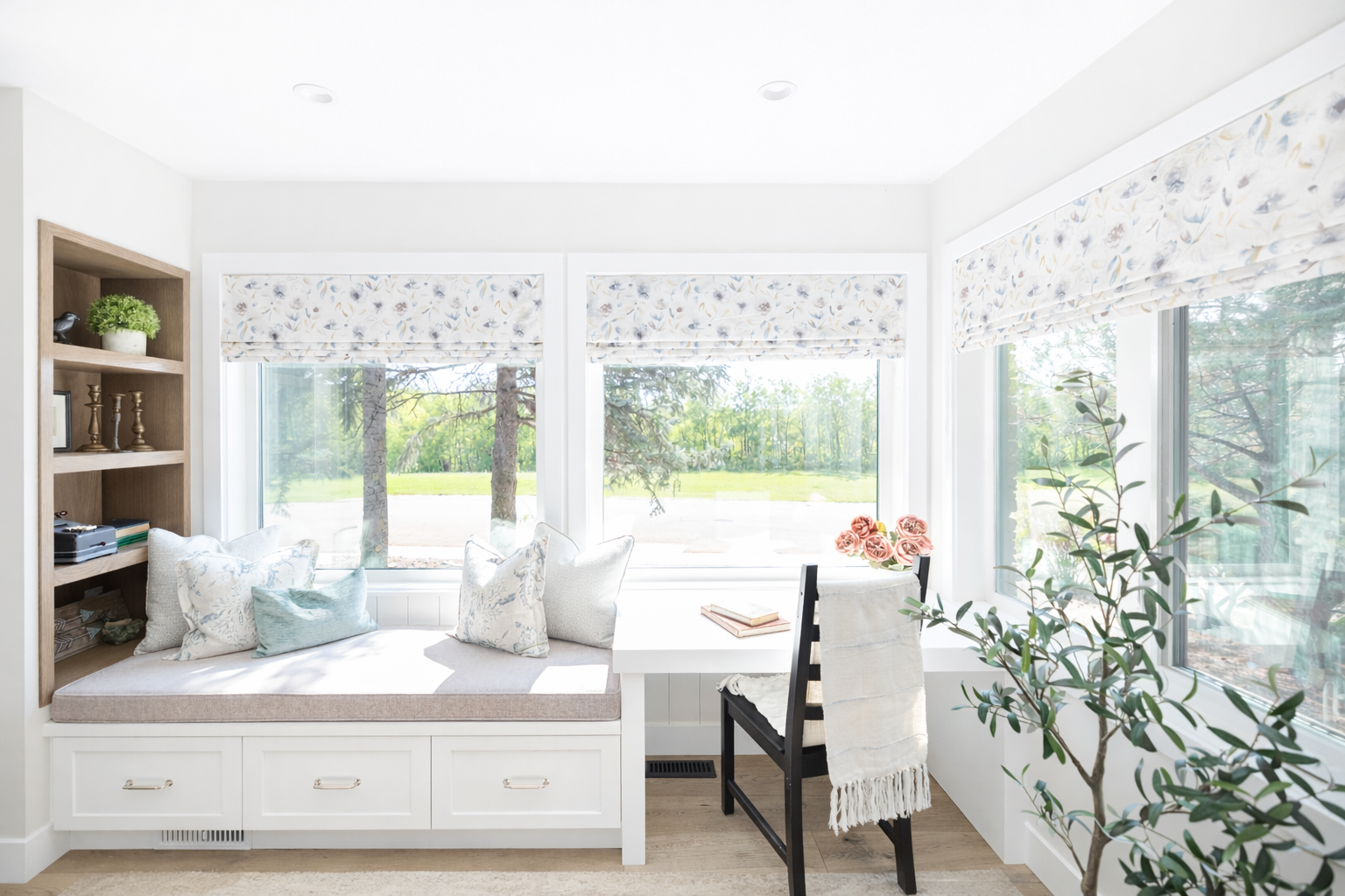 Bright sunlit nook with window seating, decorated with pillows, overlooking trees and greenery outside, with a bookshelf, plants, and books inside.