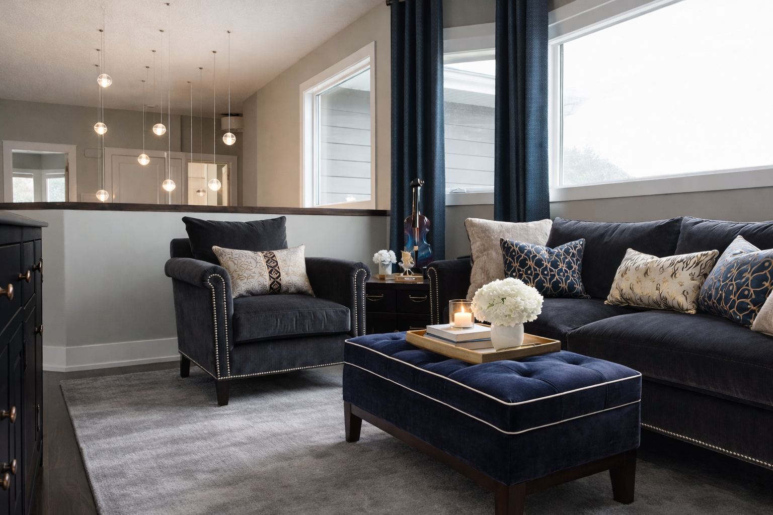 Living room with navy velvet sofa and matching armchair, decorated with patterned throw pillows, a blue ottoman with white piping, a tray with a candle and flowers, large windows with blue curtains, and a modern light fixture in the background.