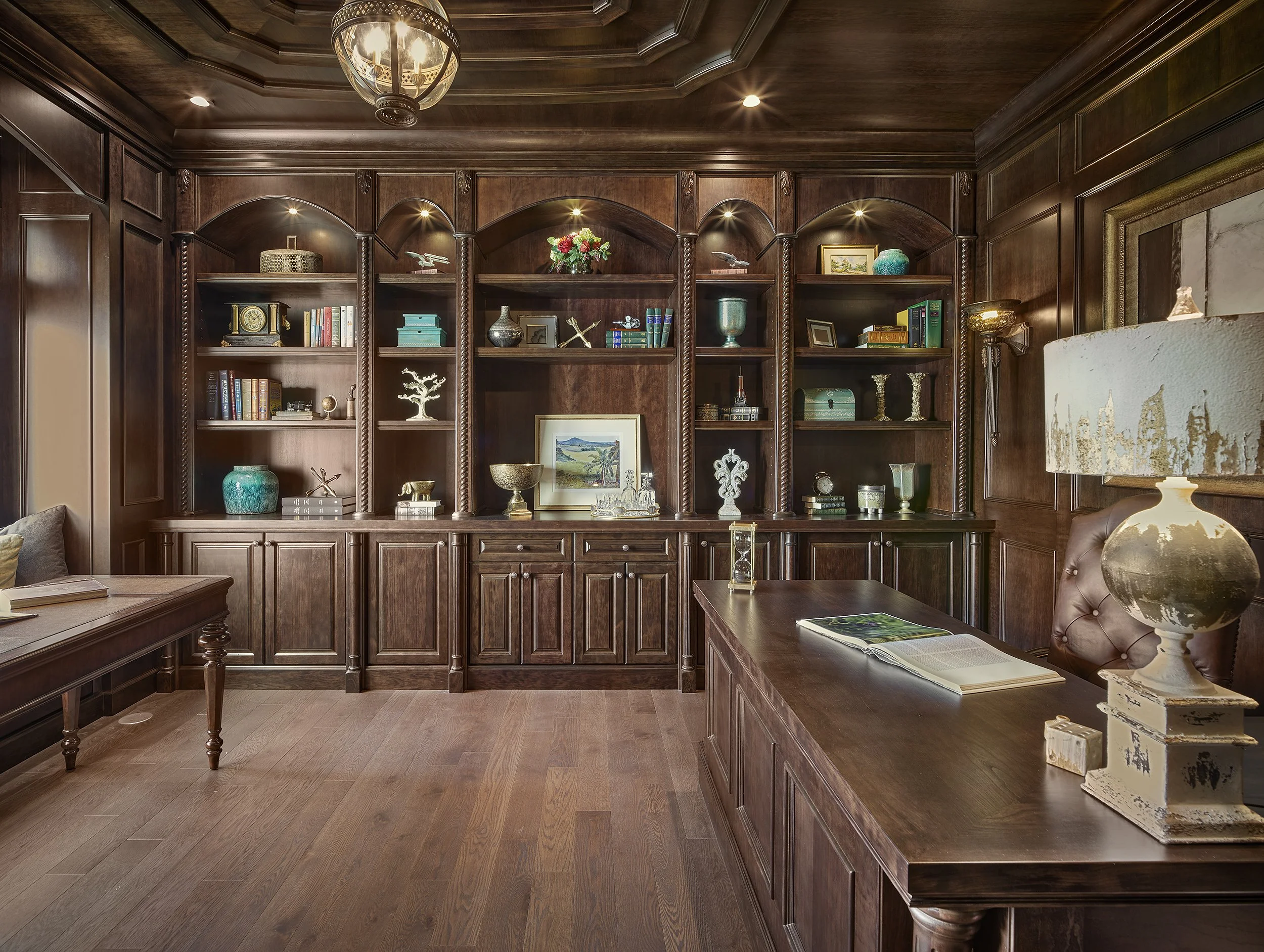 A wood-paneled office with built-in bookshelves filled with decorative items, books, and picture frames. There is a large desk with reading materials, a lamp, and a leather chair. The room has warm lighting and a wooden ceiling with a chandelier.